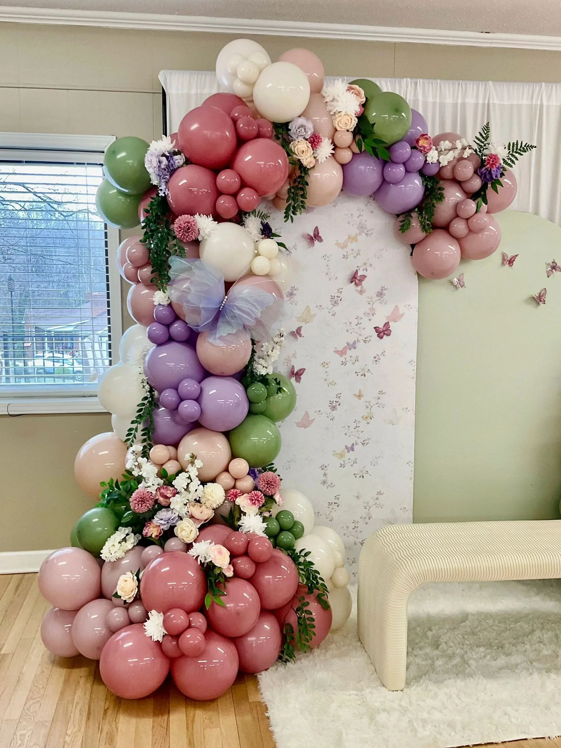 Balloon arch with pink, purple, green, cream, and white balloons, decorated with flowers and greenery, surrounding a butterfly backdrop.