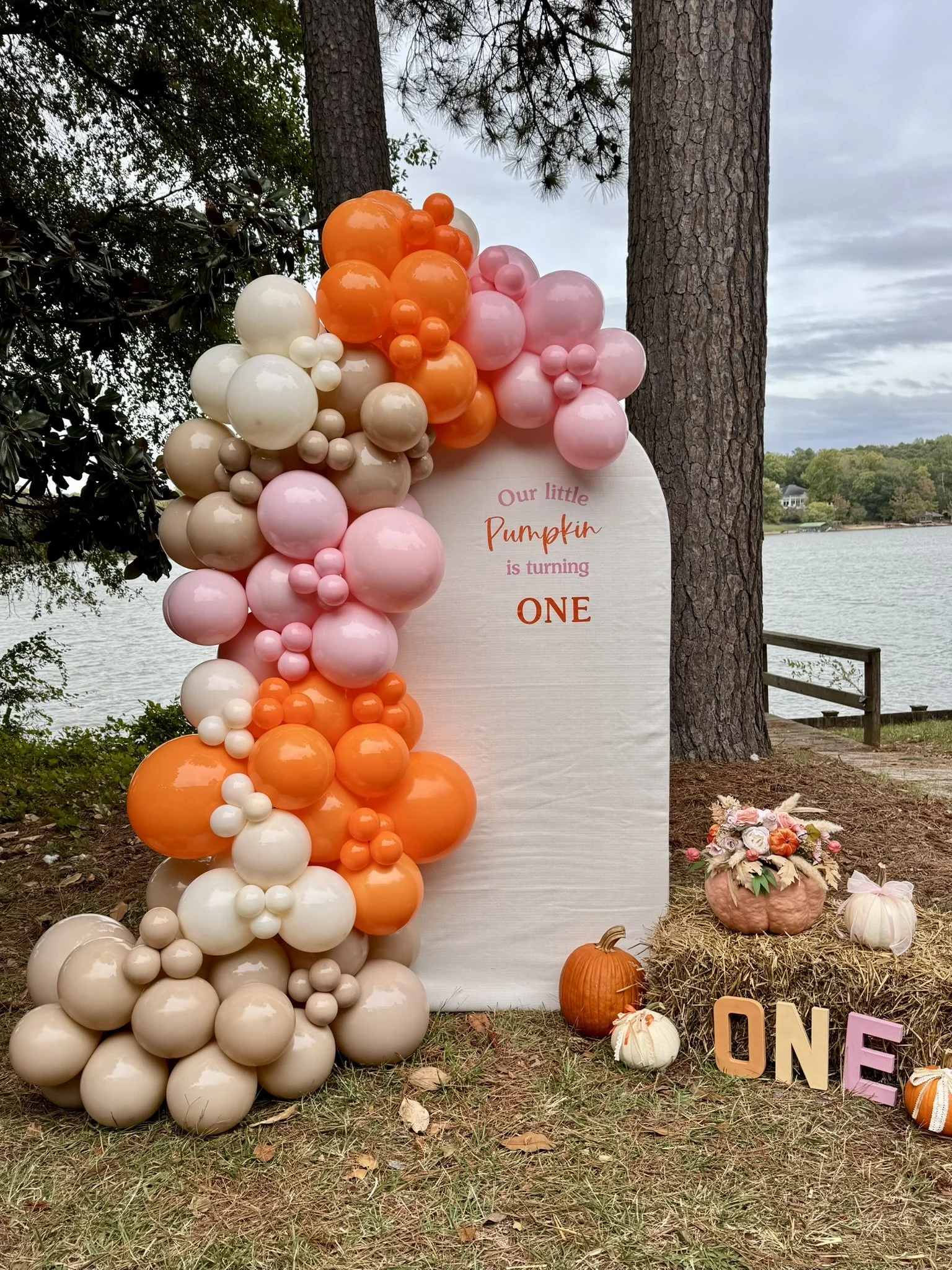 Decorative balloon arrangement with orange, pink, beige, and white balloons beside a sign that reads 'Our little Pumpkin is turning ONE.' Fall-themed pumpkins and wooden letters spelling 'ONE' are on the ground nearby, with a hay bale and flowers on 