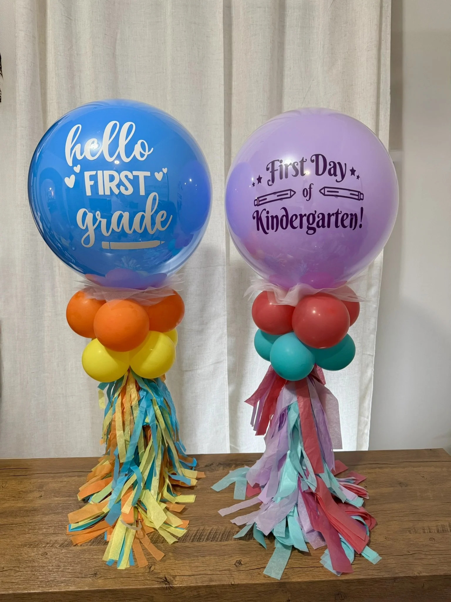 Two colorful balloons celebrating a child's first day of school, decorated with tassels and ribbons. The blue balloon on the left reads 'hello FIRST grade' and the purple balloon on the right reads 'First Day of Kindergarten!'