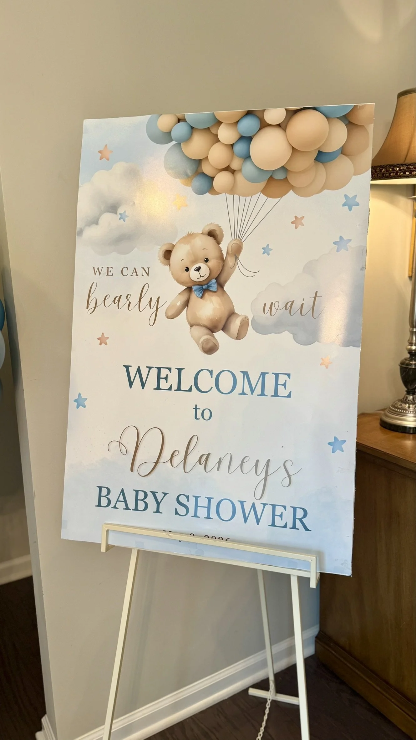 A decorative sign for a baby shower with a teddy bear holding balloons made of beige and blue spheres, surrounded by clouds and stars, welcoming guests to Delaney's baby shower.