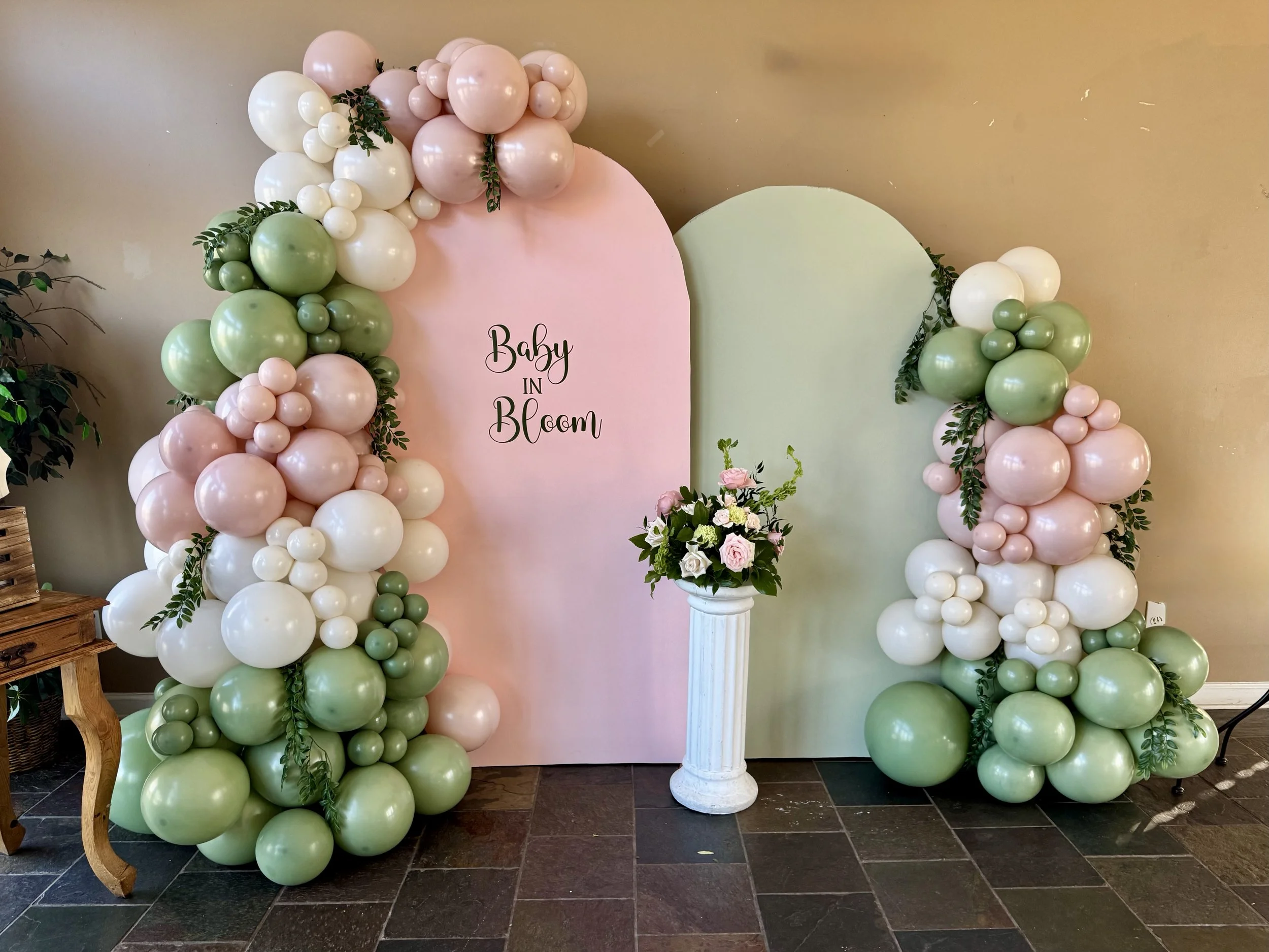 Decorative balloon arch with pastel pink, white, and green balloons, surrounding a pink and green backdrop with the phrase 'Baby in Bloom.' A white pedestal with a floral arrangement of pink and white roses and greenery is in front of the backdrop.