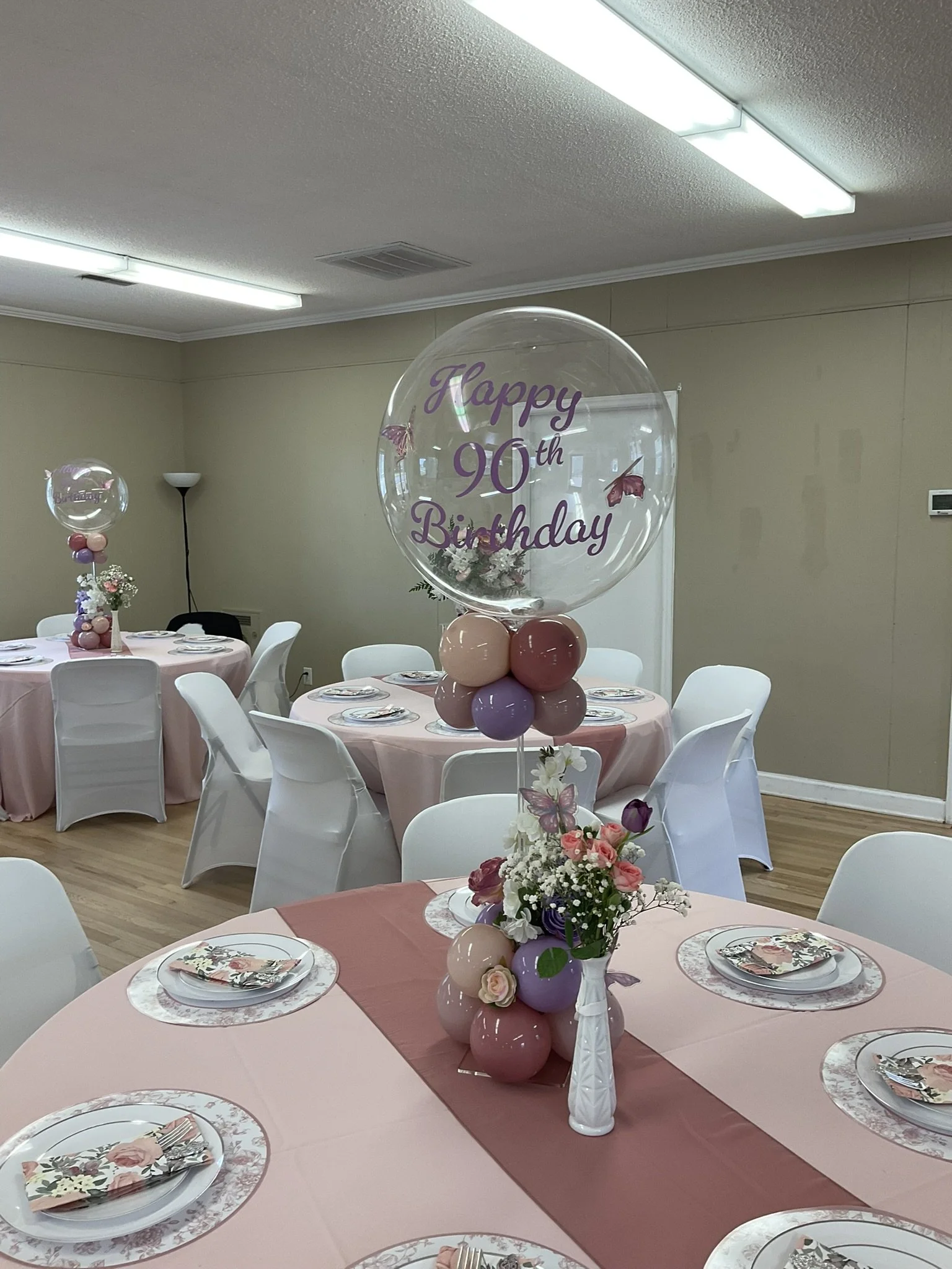 Decorated room with round tables covered in pink tablecloths for a 90th birthday celebration. Each table has floral centerpieces with pink, purple, and white flowers, and place settings with floral napkins and decorative plates. Transparent balloons with pink and purple balloons underneath, featuring a banner that reads 'Happy 90th Birthday' with butterflies, are displayed on the tables.