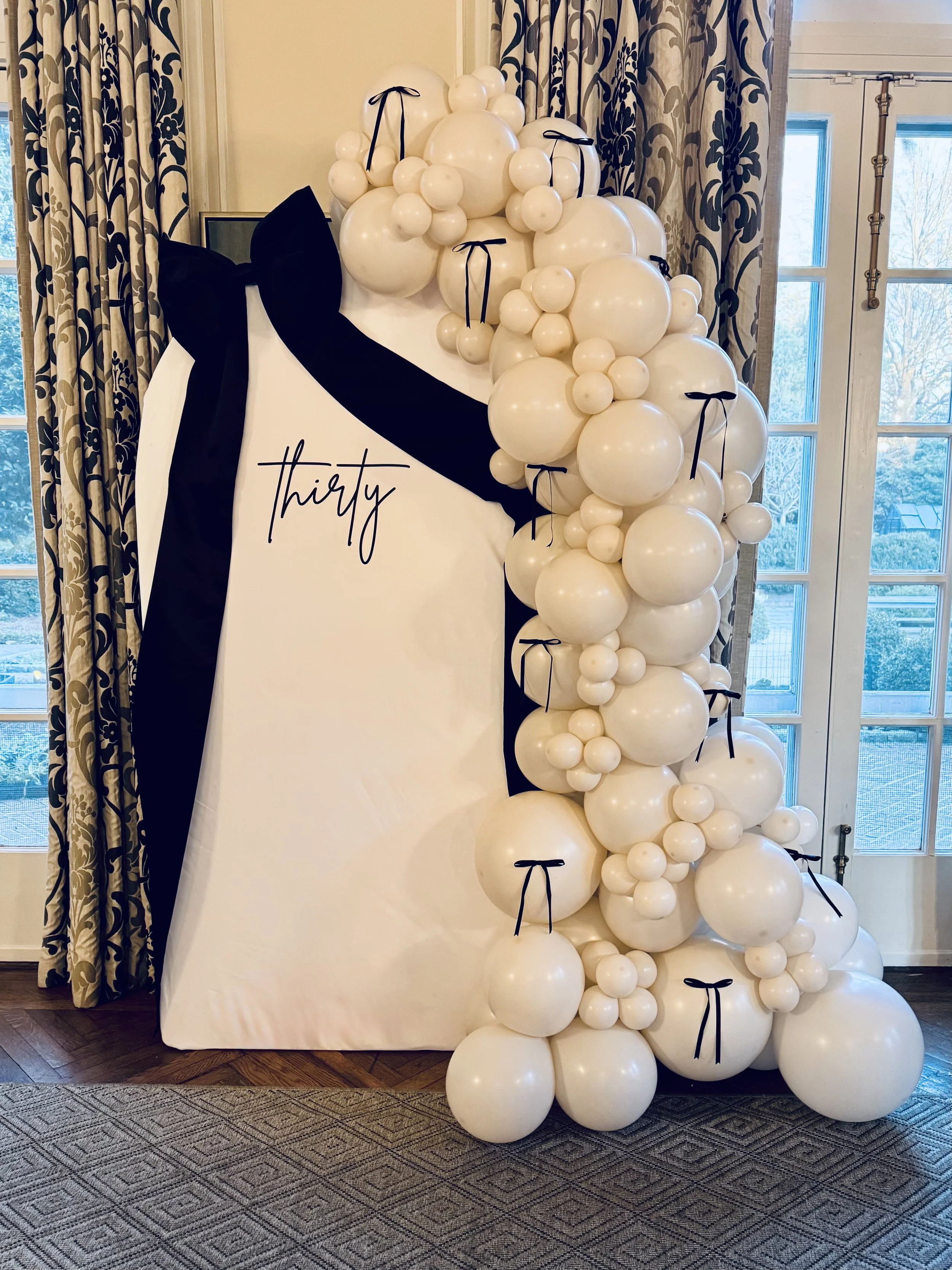 White balloon arrangement with black ribbons and a white board displaying "thirty" written in cursive, set indoors near windows with patterned curtains.