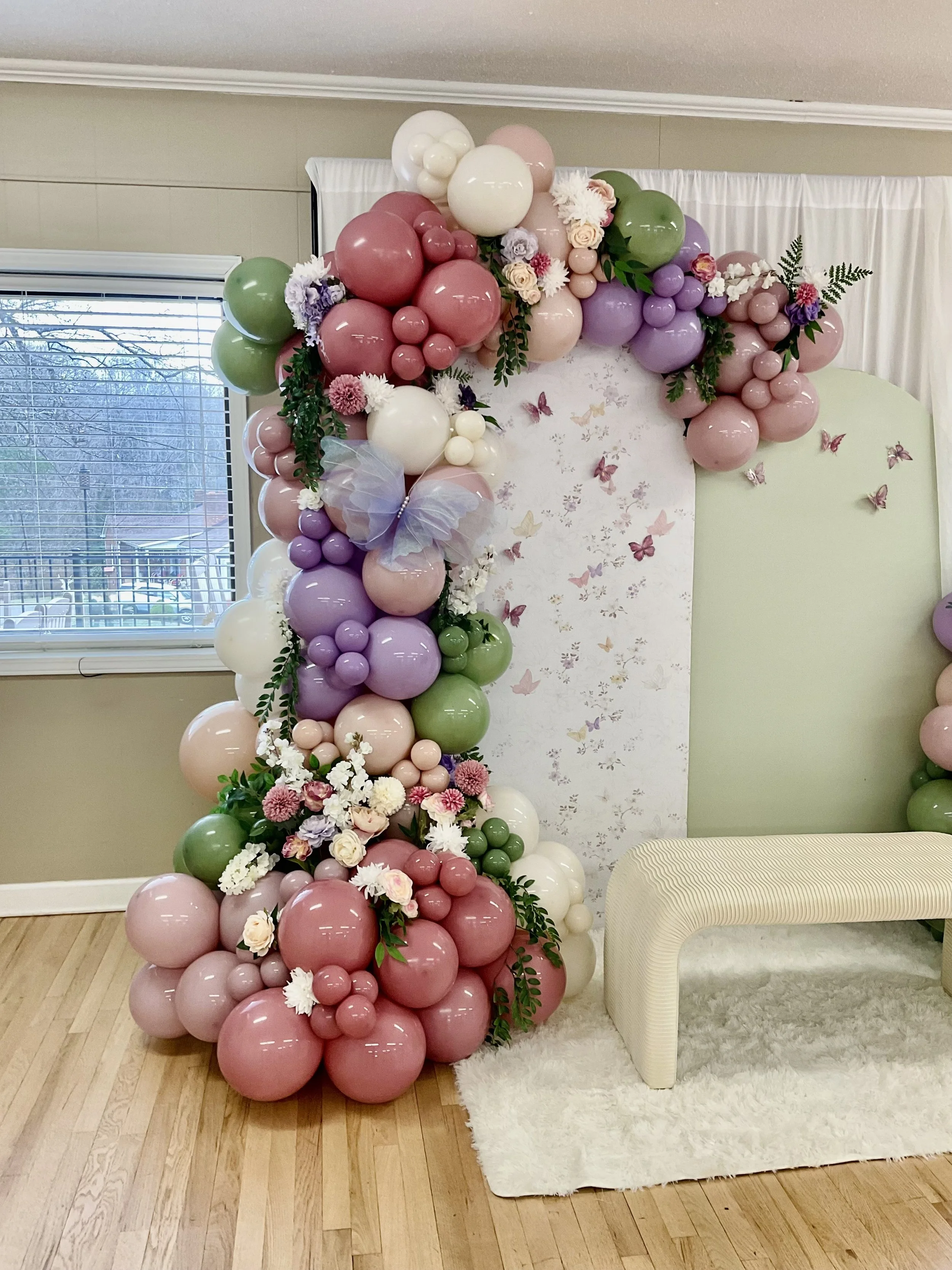 A decorative balloon arch with pink, purple, white, and green balloons decorated with flowers and greenery, set against a white curtain backdrop with butterfly wall decals, beside a window with blinds, a cream-colored bench, and a white shag rug on a