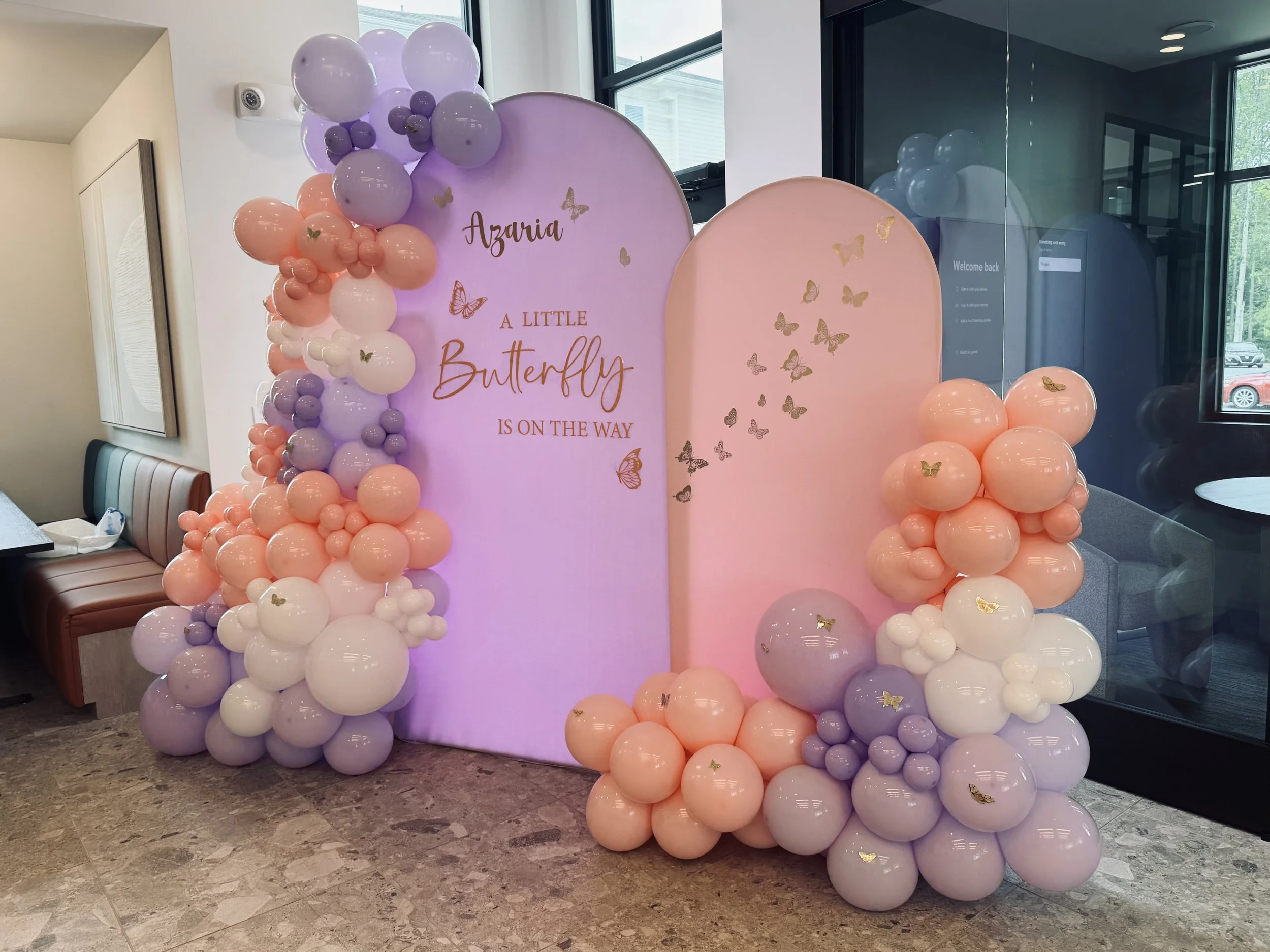 Decorative backdrop for a celebration with pastel pink, purple, and white balloons arranged around three large oval panels. The left panel has the words "LITTLE BUTTERFLY IS ON THE WAY" written on it, and the right panel is decorated with butterfly s