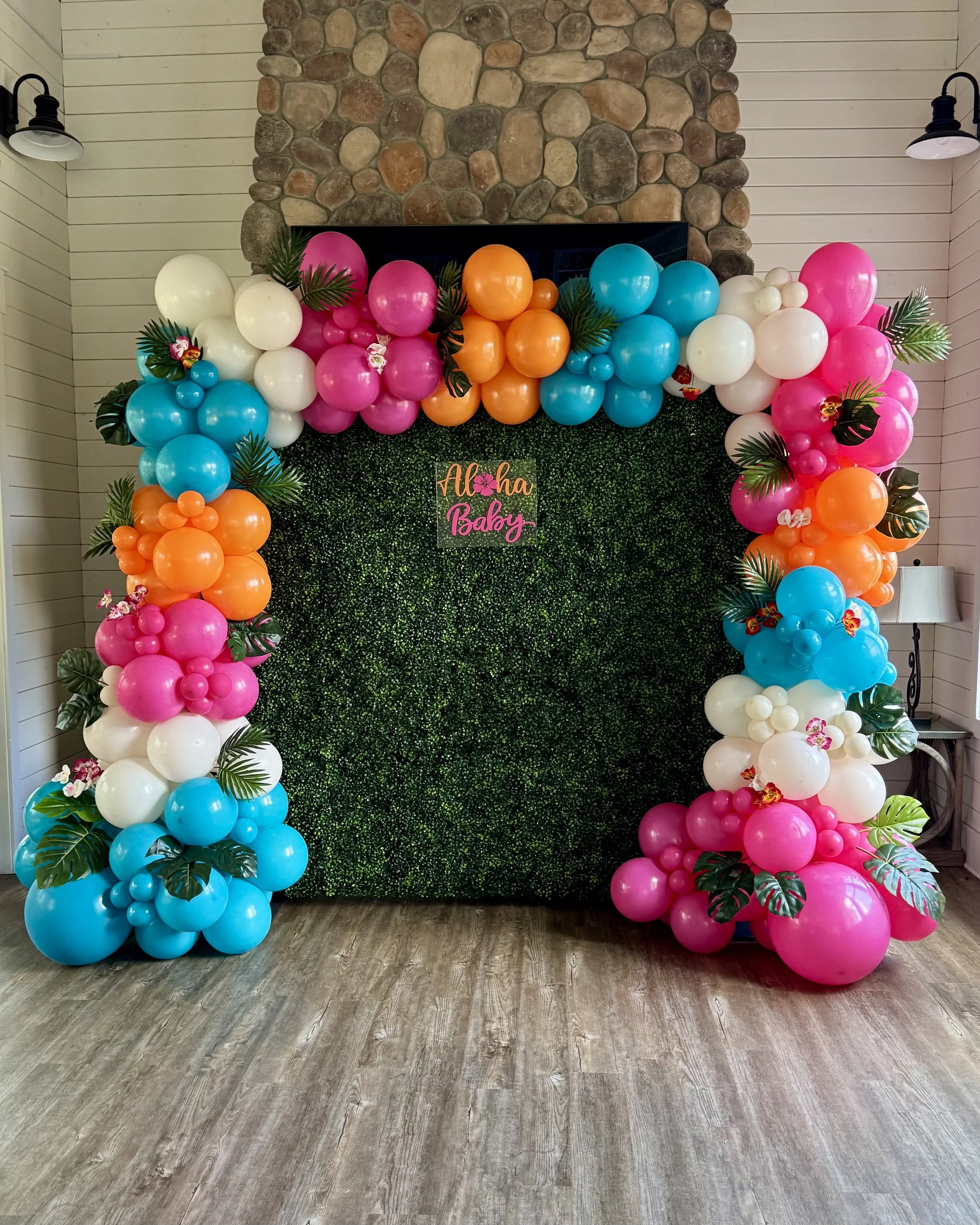 Colorful balloon arch with pink, orange, blue, and white balloons, decorated with tropical leaves and orchids, in front of a faux green hedge backdrop and a stone fireplace.