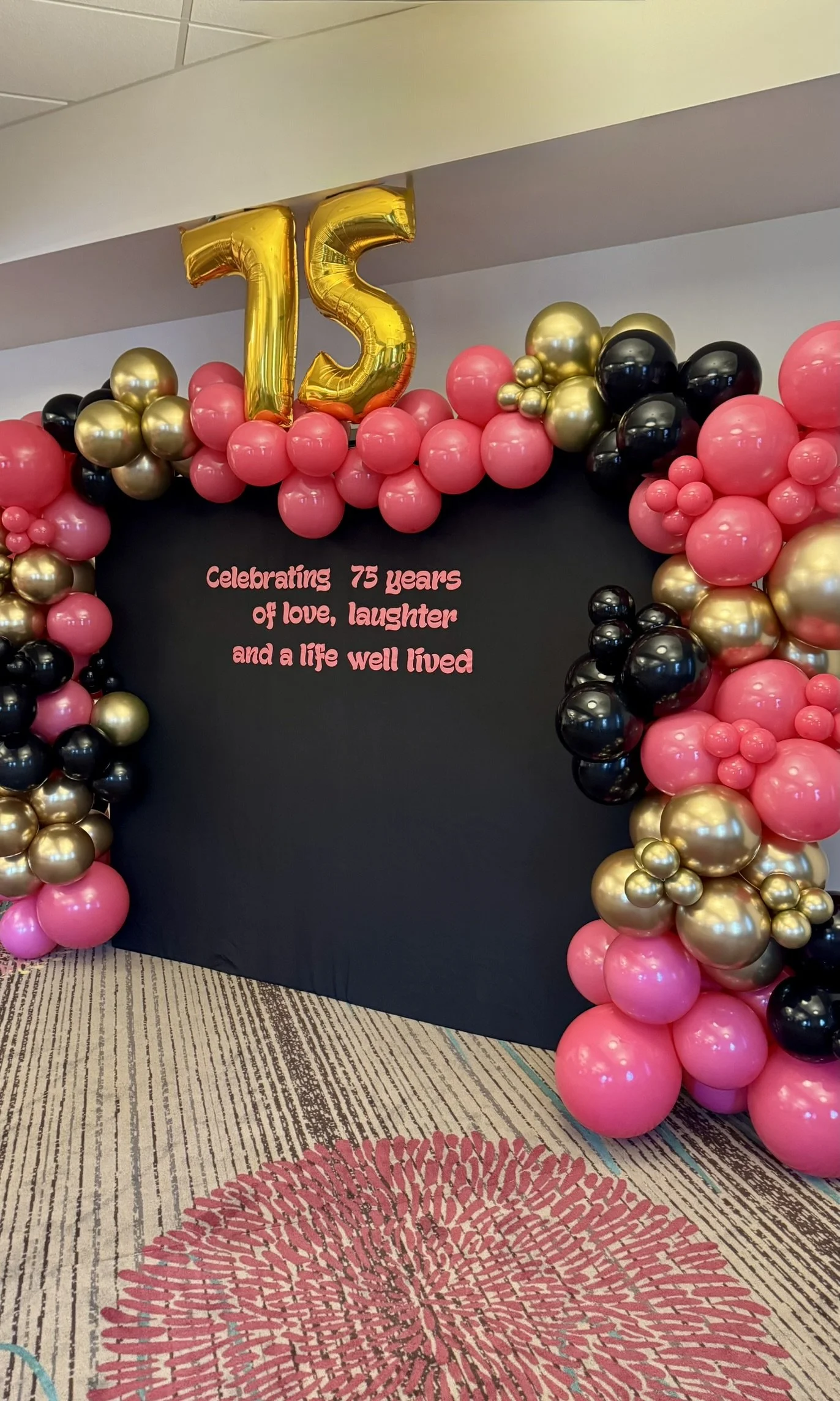 Balloon arch with pink, gold, and black balloons, gold number balloons forming '75', and a black backdrop with pink lettering celebrating 75 years with the message: 'Celebrating 75 years of love, laughter and a life well lived.'
