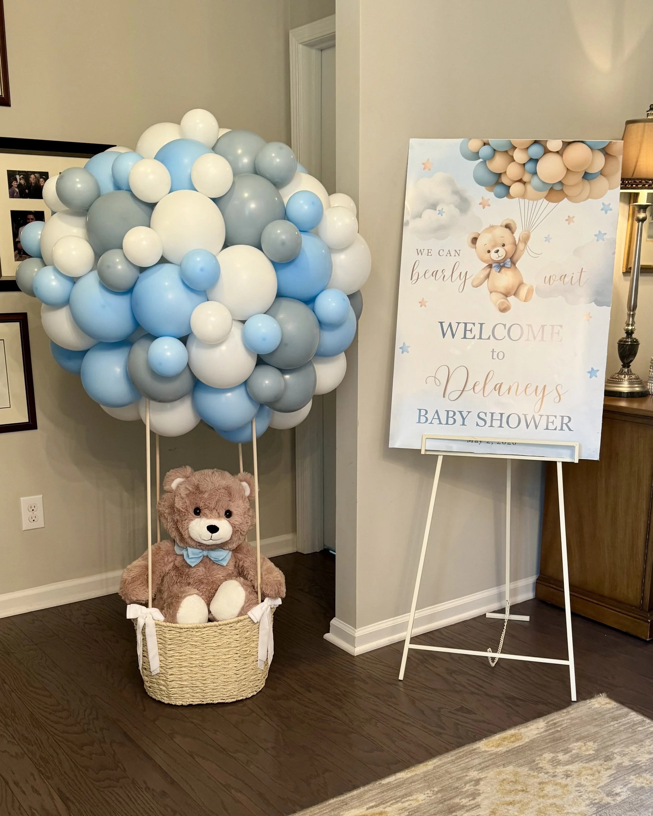 Decorative display for a baby shower, featuring a large cluster of blue, gray, and white balloons, a stuffed teddy bear with a blue bow sitting in a woven basket, and a welcome sign with a teddy bear holding balloons, clouds, and stars.