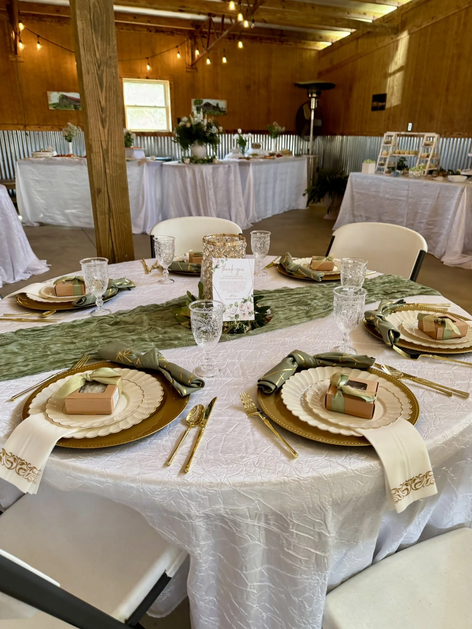 Round table with white tablecloth, gold chargers, elegant napkins, glasses, and small gift boxes, set for a celebration or wedding reception in a rustic decorated venue with string lights and wooden walls.