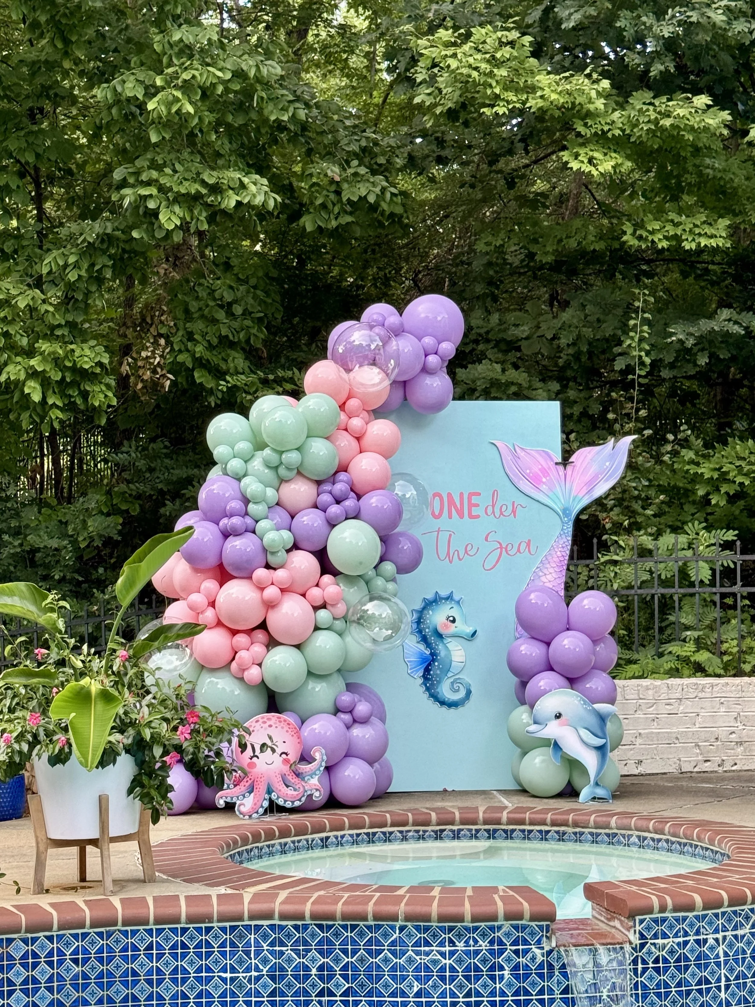 Decorative mermaid-themed party backdrop with pastel balloons, illustrated sea creatures, and the phrase 'One-der the Sea' near a small pool.