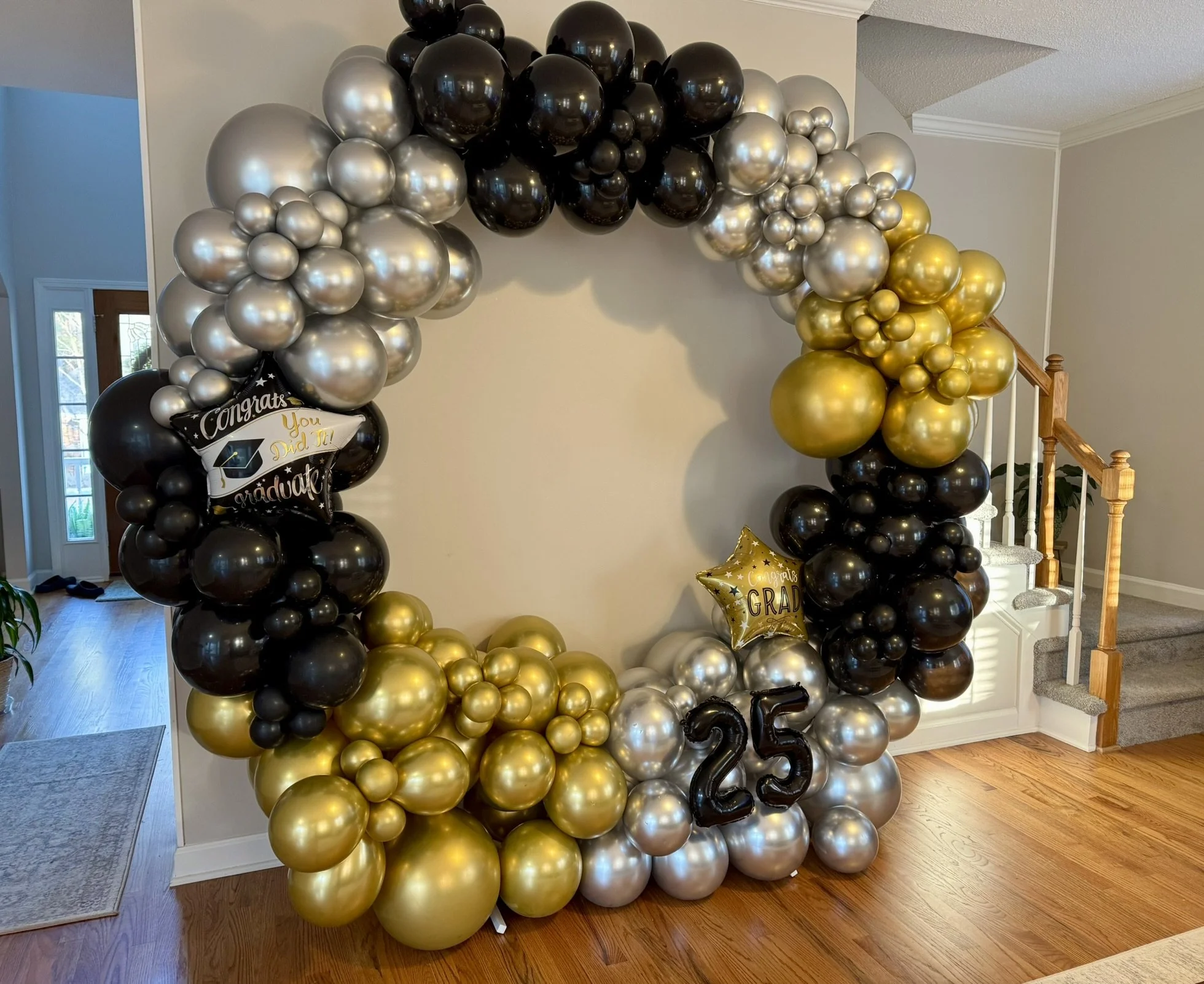A large graduation balloon arch in gold, silver, and black colors, set up indoors. The arch features a congratulatory message with balloons spelling out "25" and a star-shaped balloon with "Congrats Grad" written on it.
