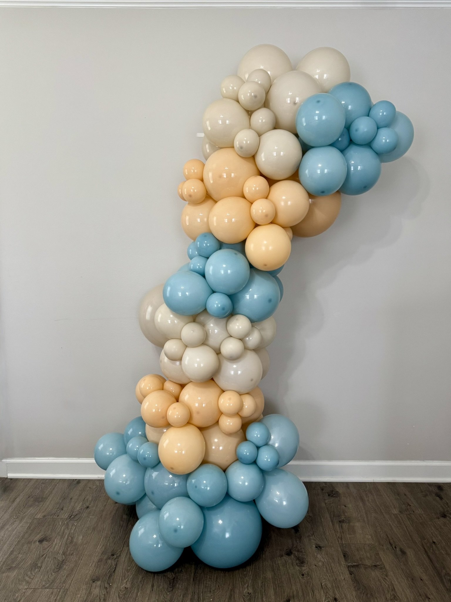 Cluster of pastel-colored balloons in shades of white, cream, peach, and blue arranged in a vertical, abstract sculpture against a plain wall.