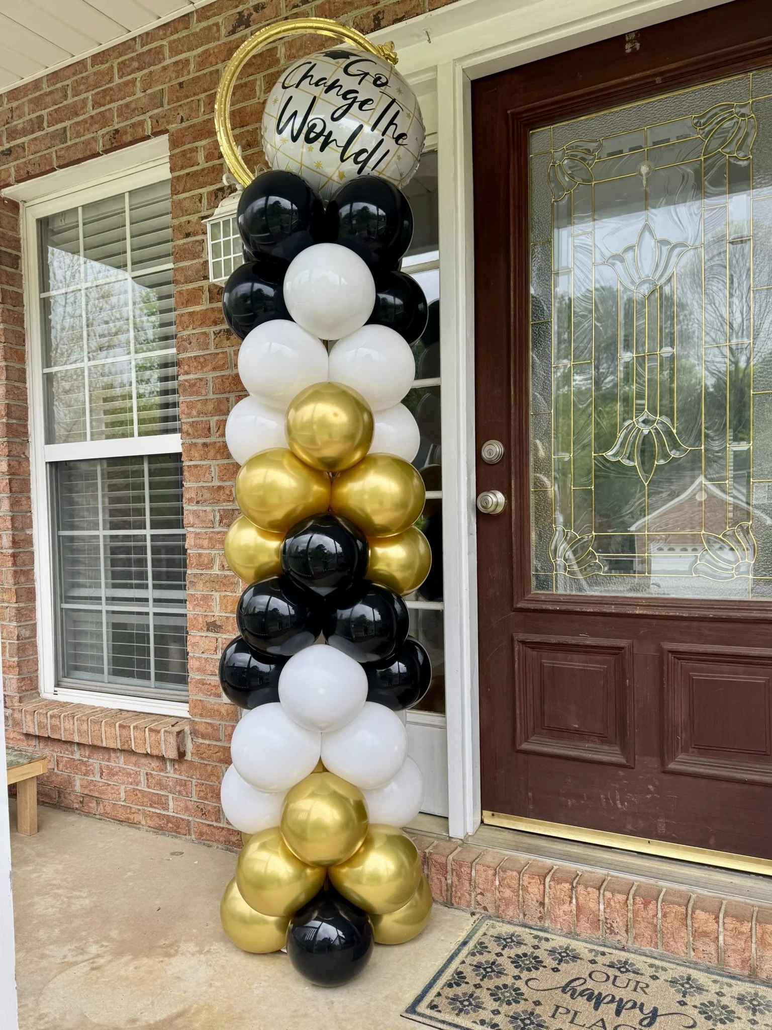 Graduation balloon column with black, white, and gold balloons topped by "Go Change the World" balloon. Outside at the porch door entrance.