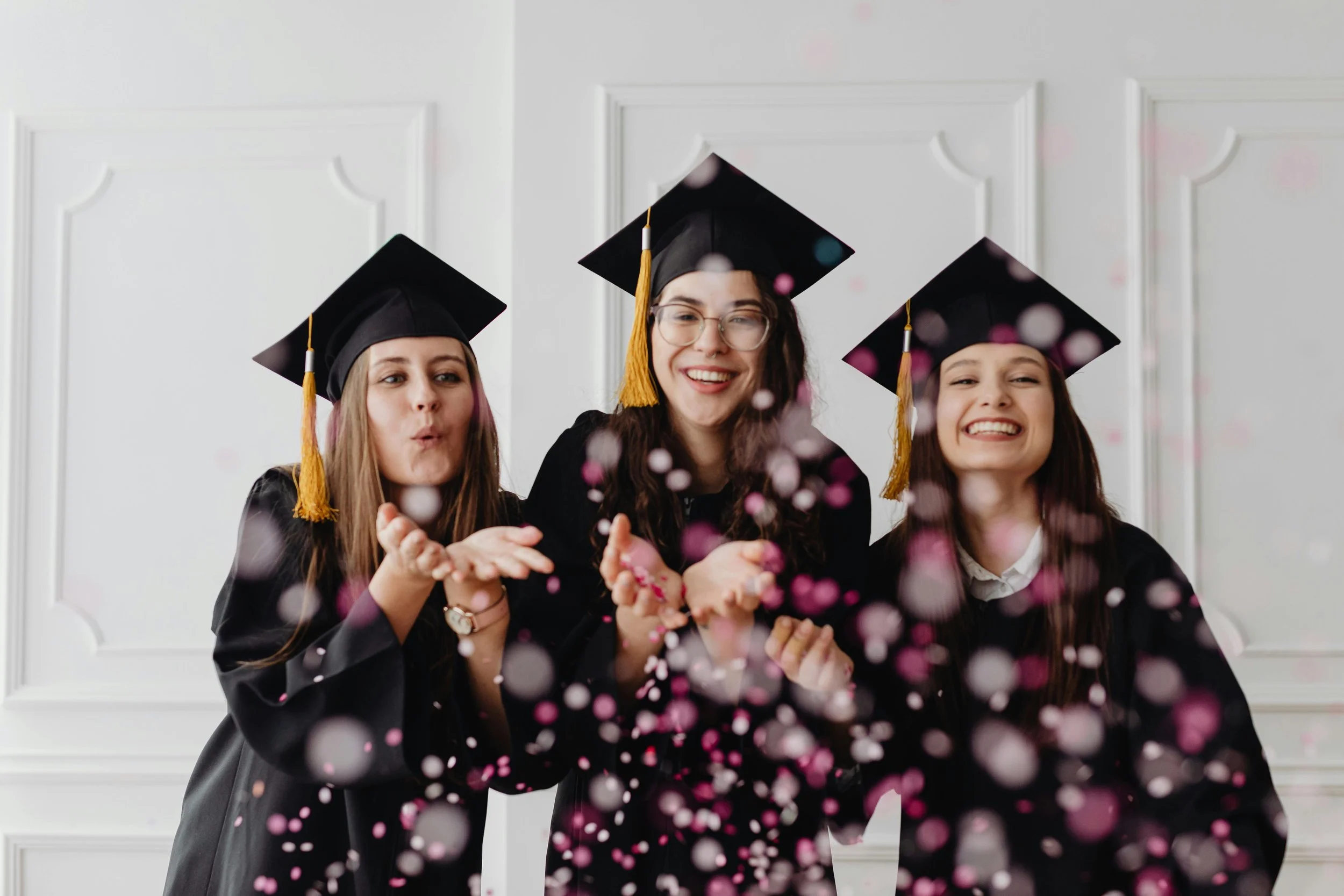 Three graduates wearing caps and gowns, smiling and throwing confetti.