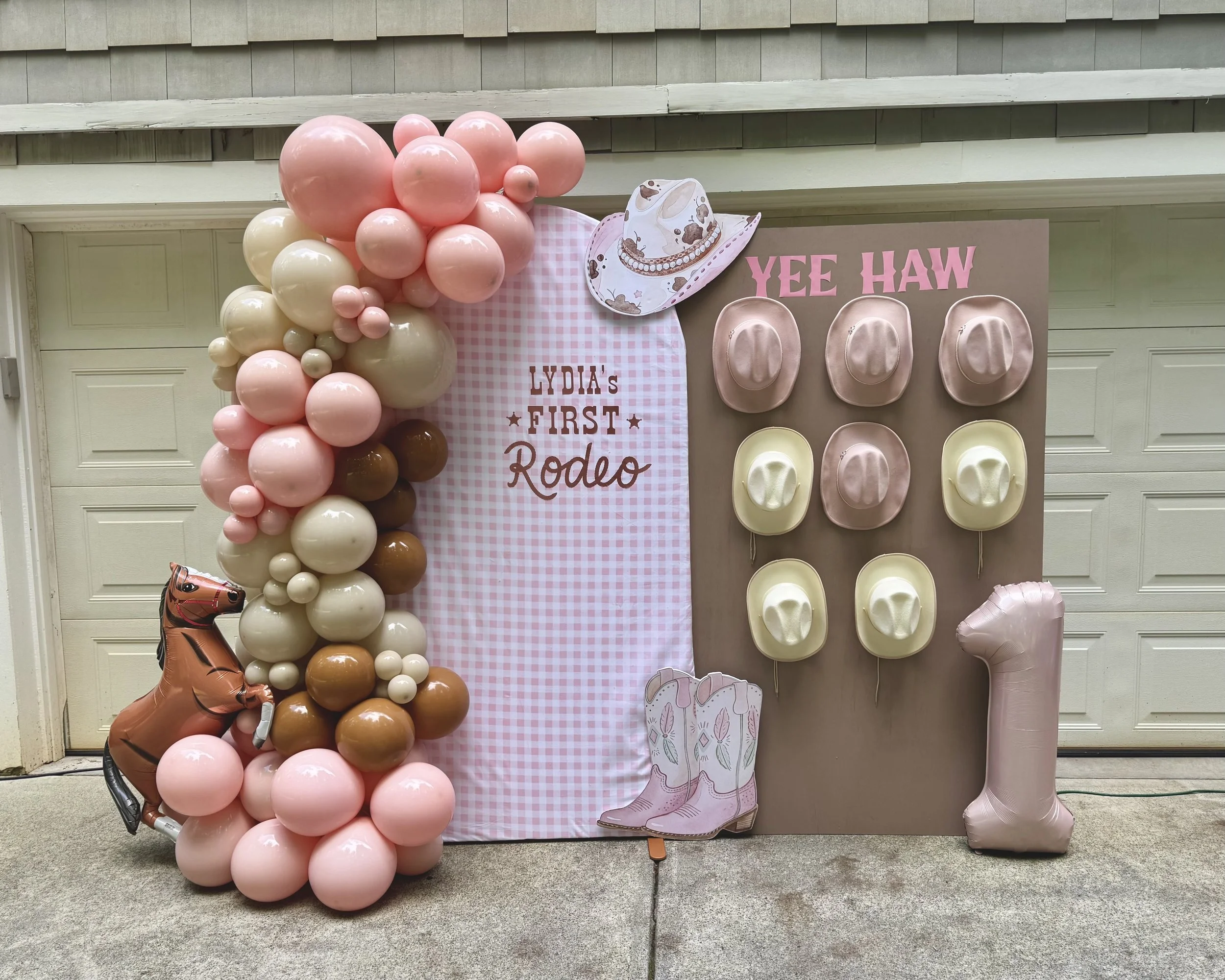 Decorative backdrop for a birthday celebration with pink, white, and brown balloons, a paper cowboy hat, pink cowboy boots cutout, and a large pink number one balloon. The backdrop features text that reads "First Rodeo" and "Yee Haw".