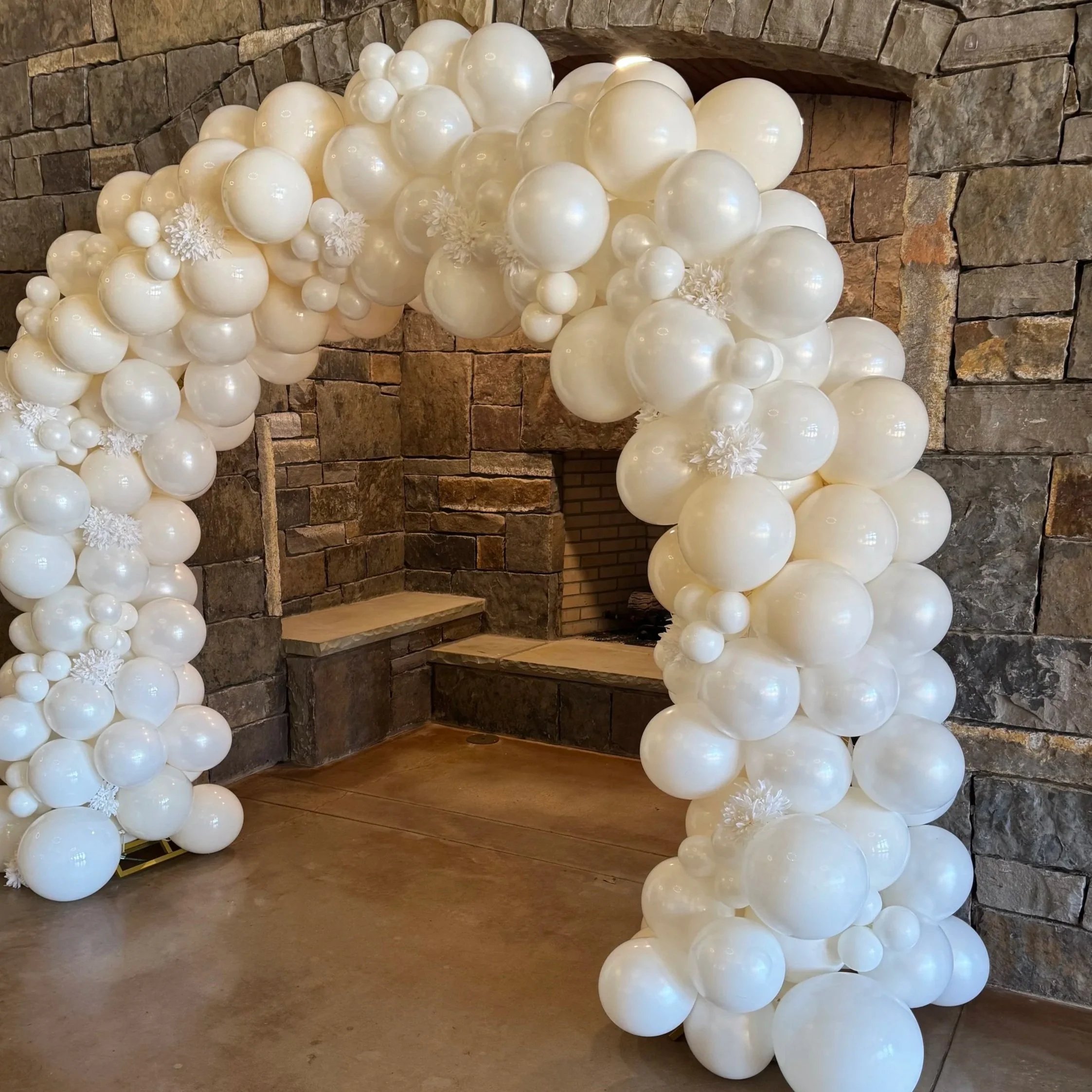 A balloon arch made of white balloons of various sizes, decorated with snowflake ornaments, set against a stone wall in an indoor setting.