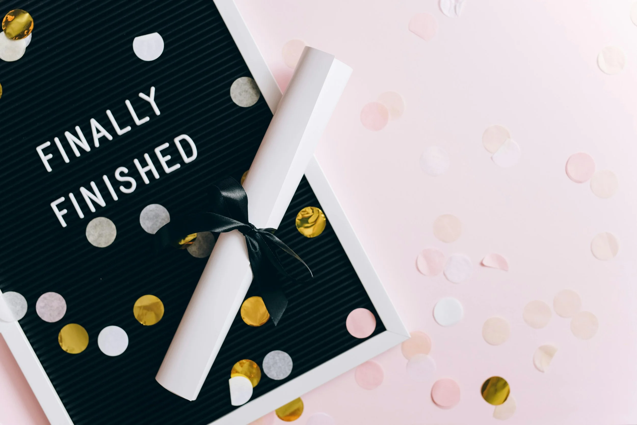 Letter board with 'Finally Finished' message, diploma with black ribbon, and confetti on pink background.