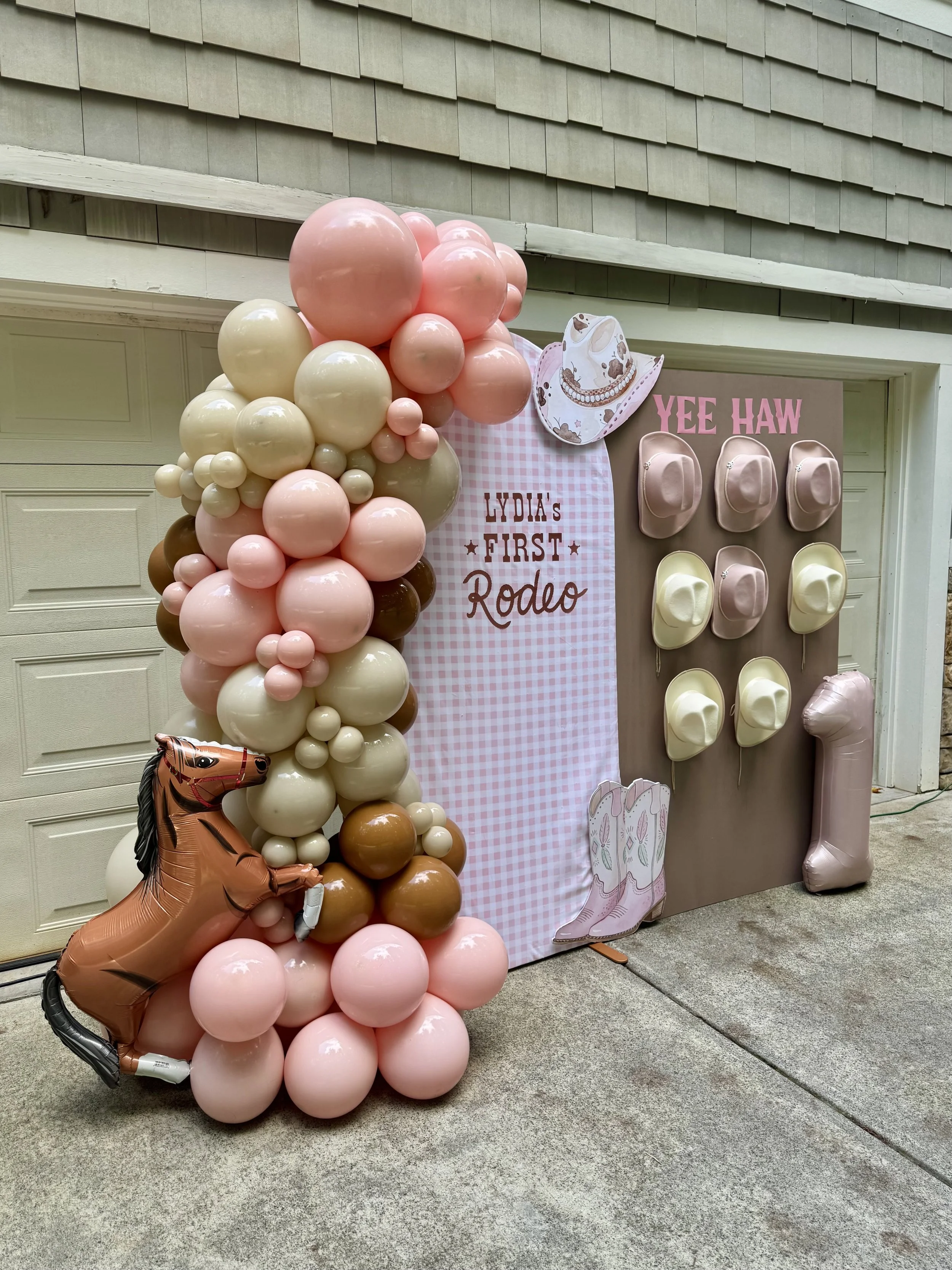 Decorative display for first rodeo birthday featuring a balloon arch with pink, white, brown, and cream balloons, a backdrop with a gingham pattern and cowboy theme, pink hats, a cowboy boot cutout, a horse balloon, and a pink hat-shaped balloon.