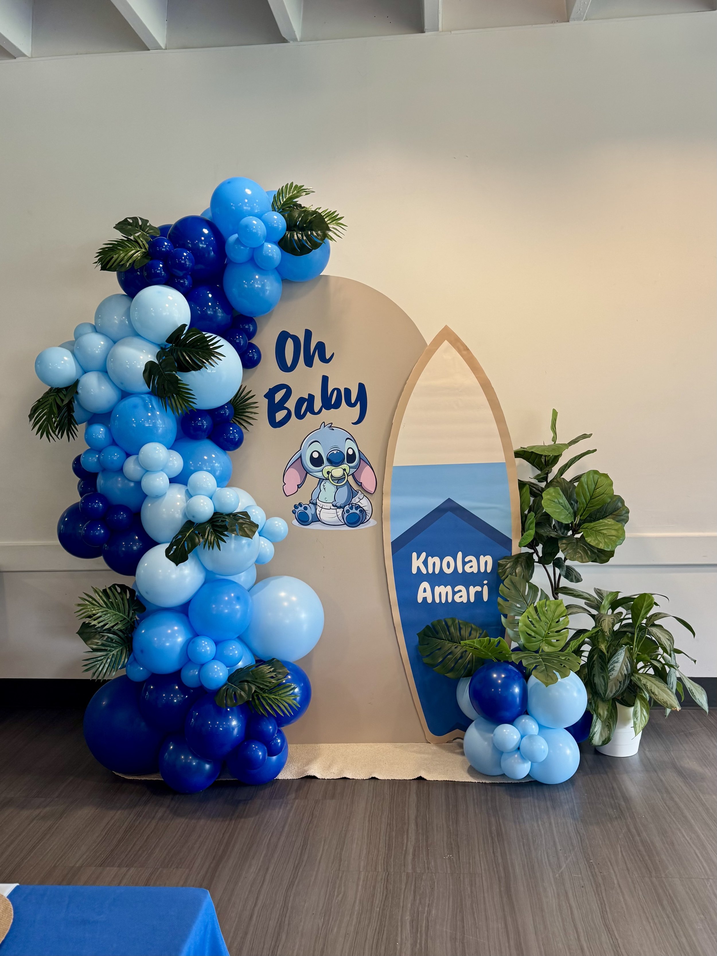 Baby Stitch shower decoration with a balloon arch in various shades of royal blue, featuring Baby Stitch, and a backdrop with the words 'Oh Baby' on a surfboard-shaped print, complemented by green tropical plants.