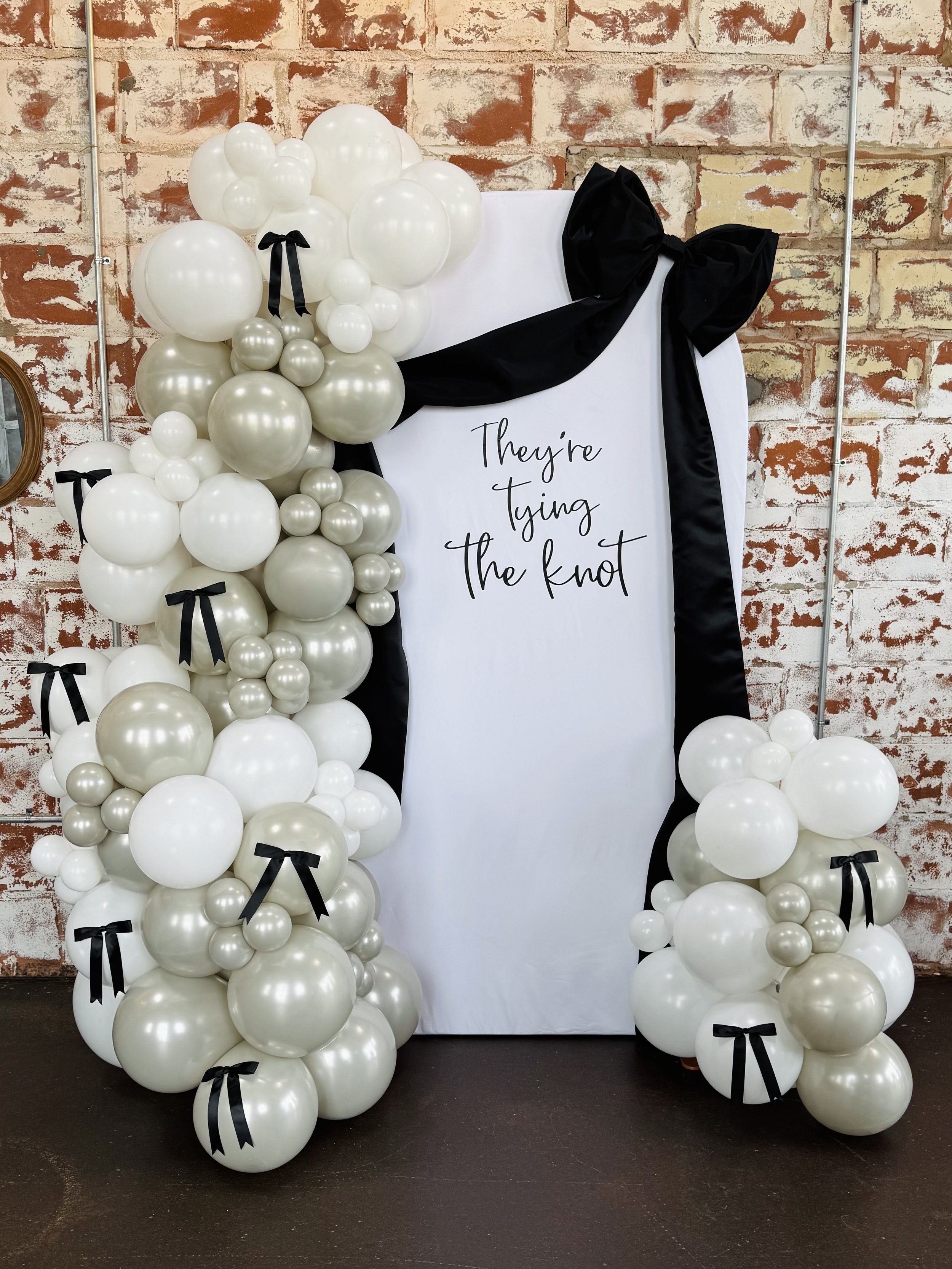 Balloon garland on an arch frame made of white and pearl-colored balloons with black ribbon bows, positioned around a white backdrop with the text 'They're Tying the Knot' written on it.