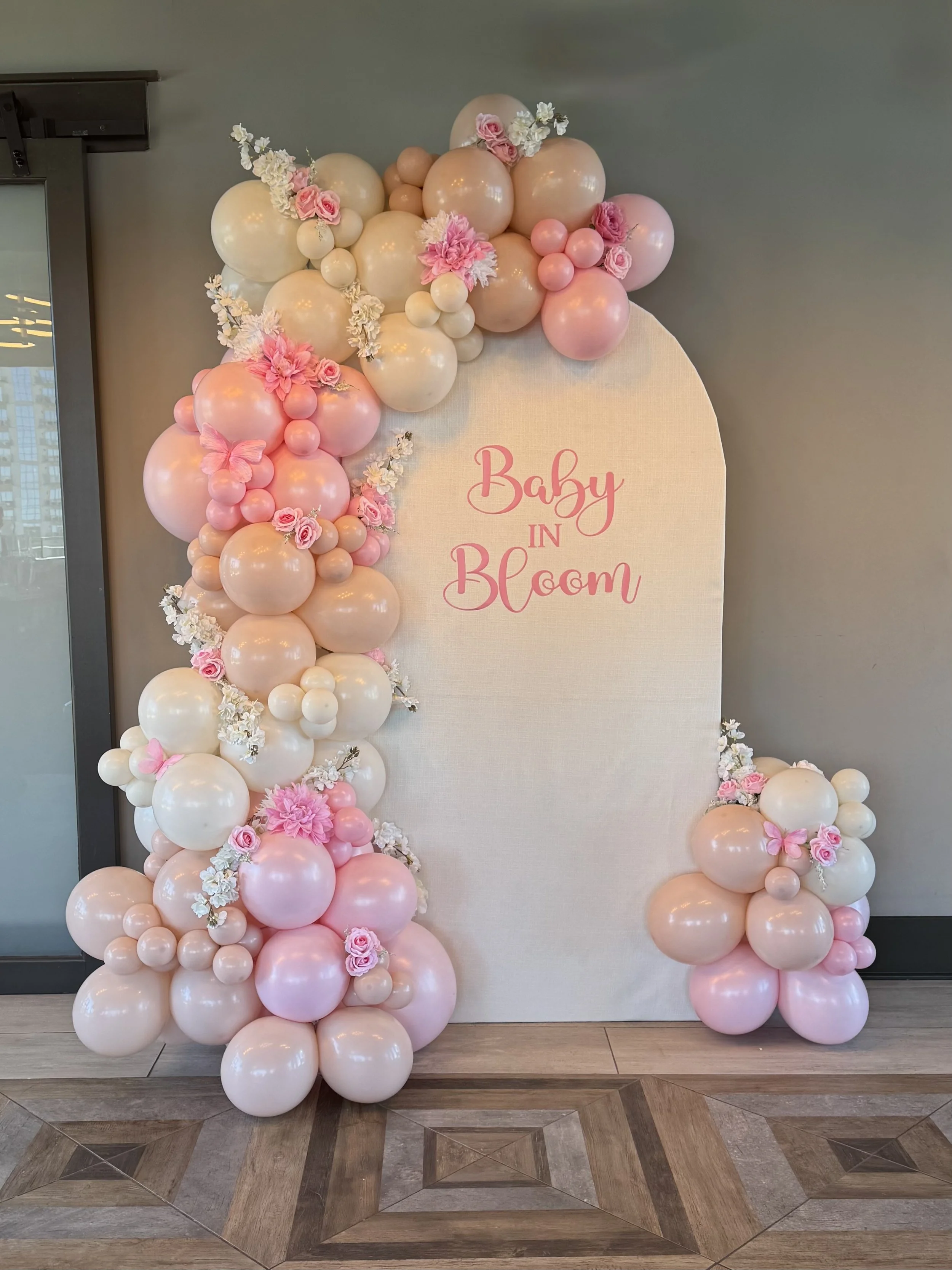 Decorative arch made of pink, white, and peach balloons with flowers, with a white backdrop that has pink cursive text reading 'Baby in Bloom'.
