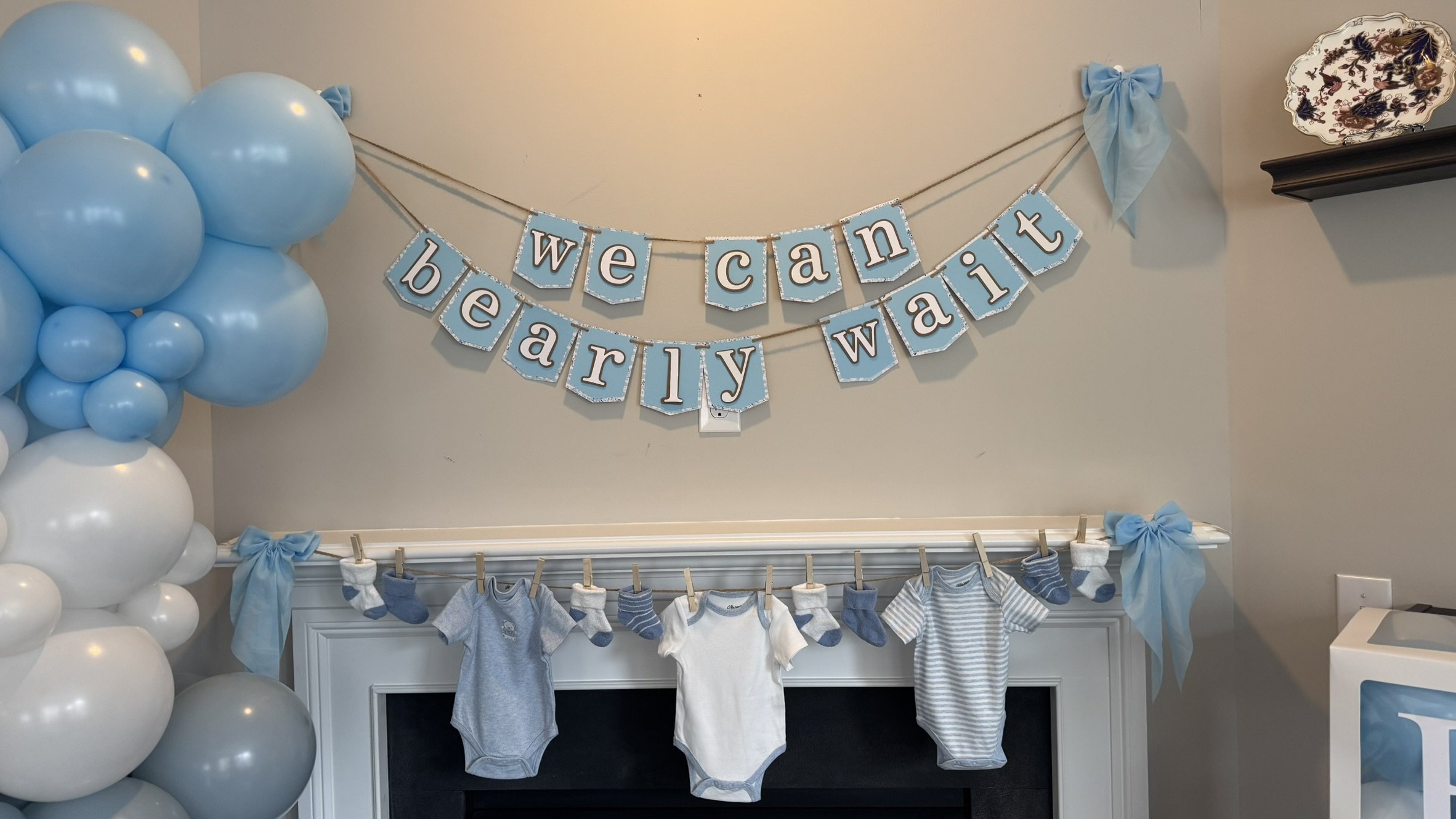 Baby shower decor with a blue and white theme, featuring a balloon arrangement on the left, a "we can't what" banner on the wall, and a white mantel with hanging baby clothes and socks.