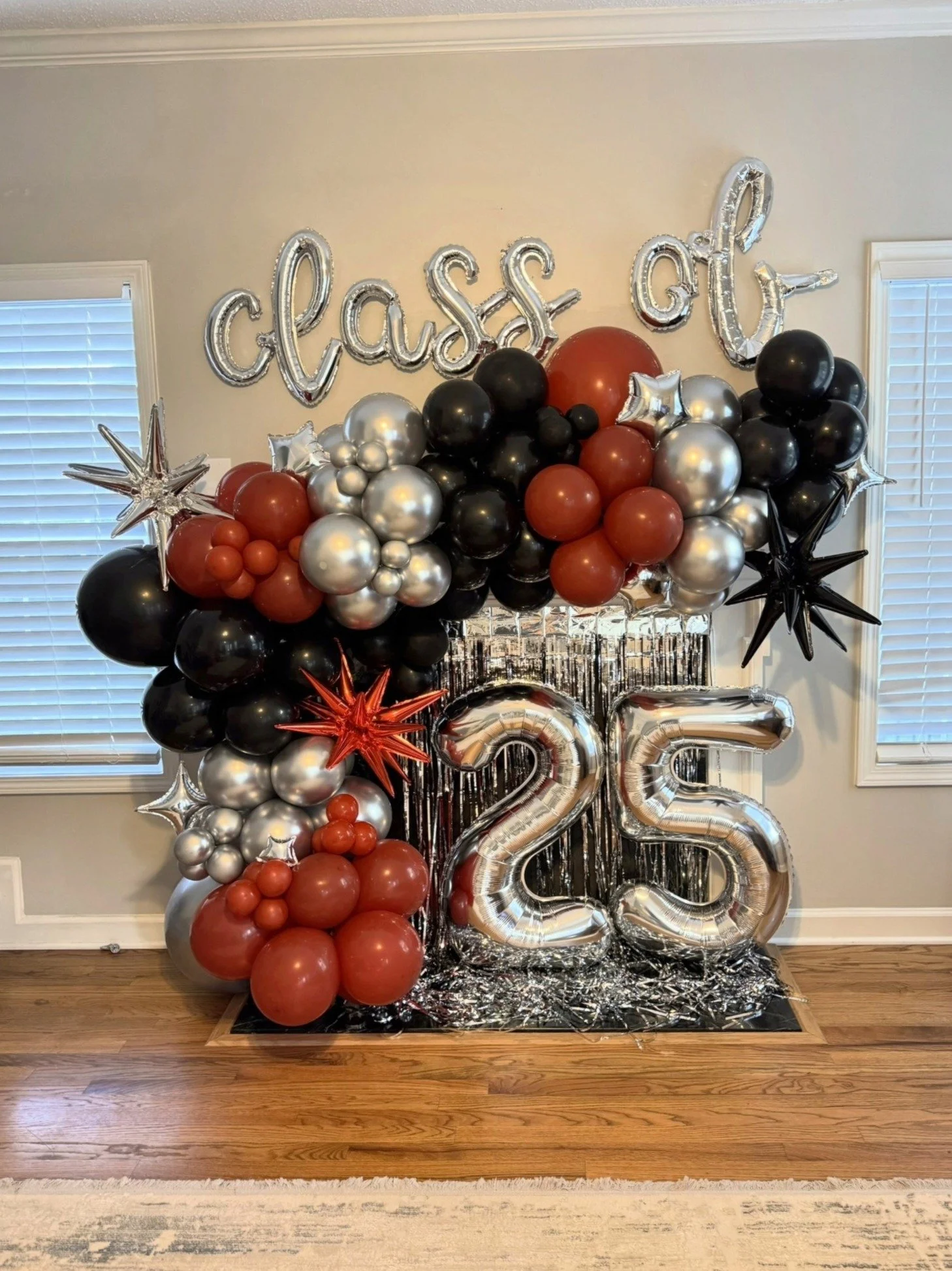 Graduation balloon display with "class of 25" in metallic balloons, surrounded by red, black, and silver balloons, with silver fringe background.