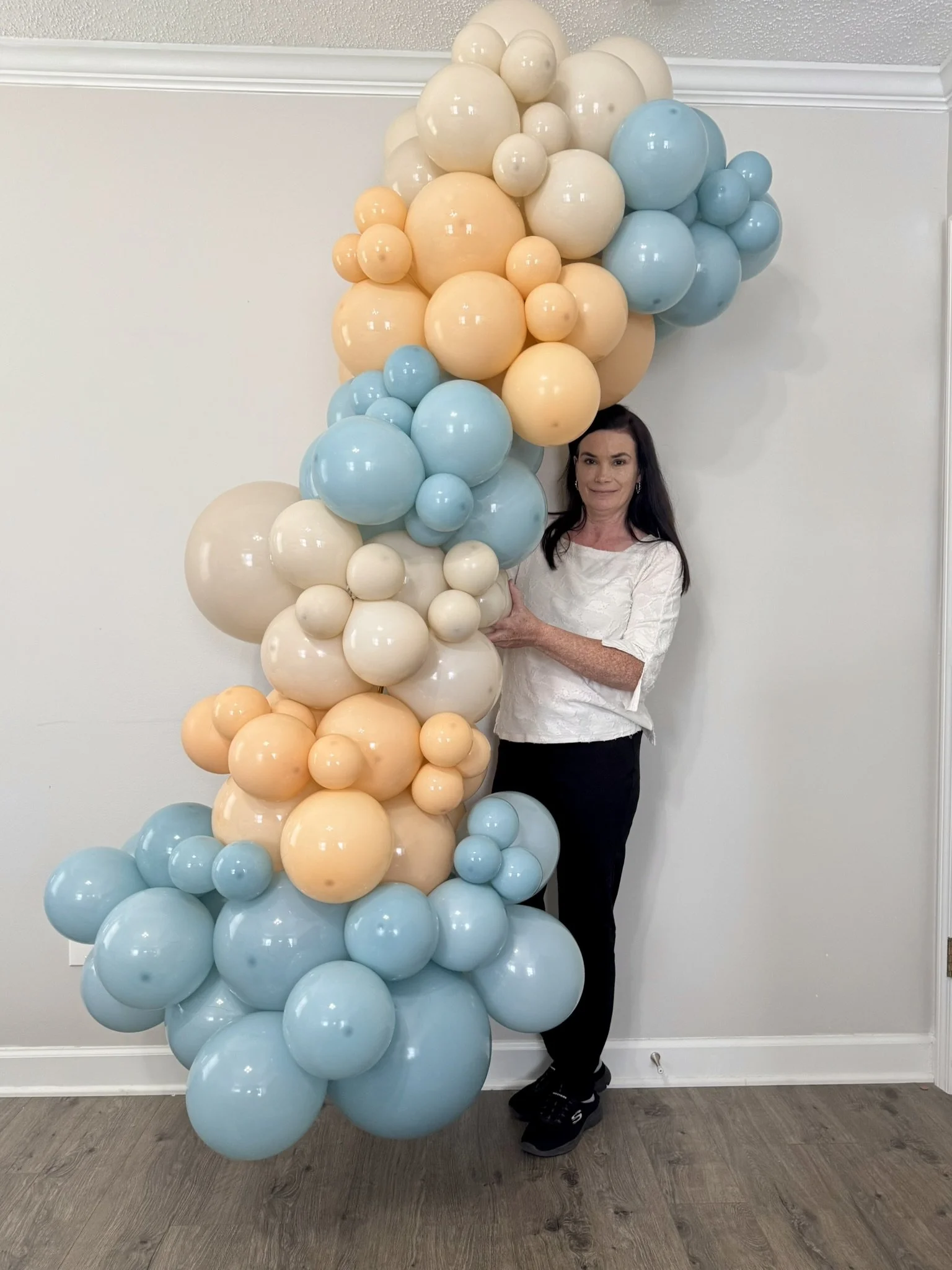 Woman standing next to a large cluster of pastel-colored balloons in shades of blue, peach, and cream.
