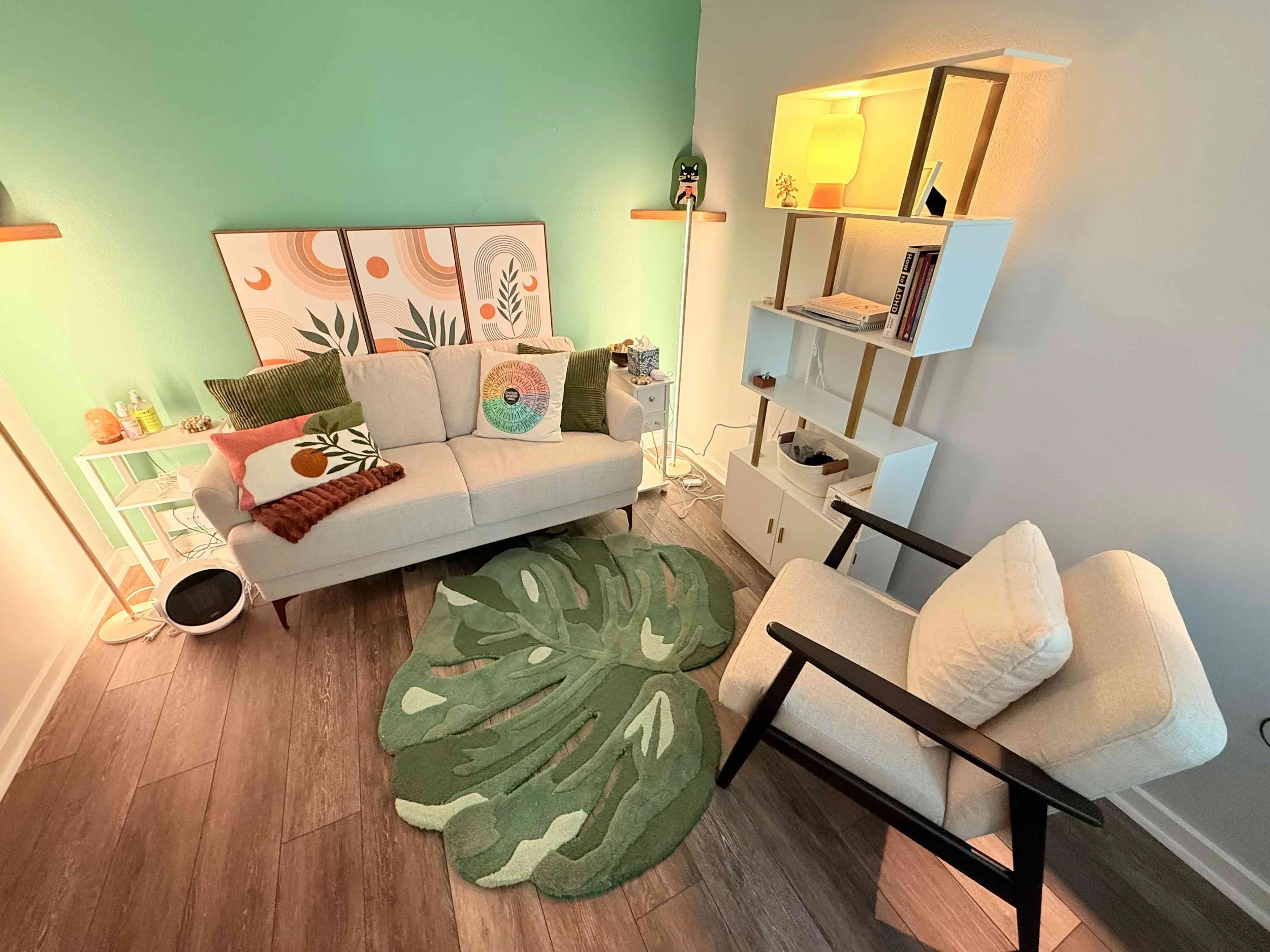 Living room with a white sofa, throw pillows, a green leafy rug, a white armchair, and a modern shelving unit with books, a lamp, and decorative items, wooden flooring, and green and neutral-colored walls.