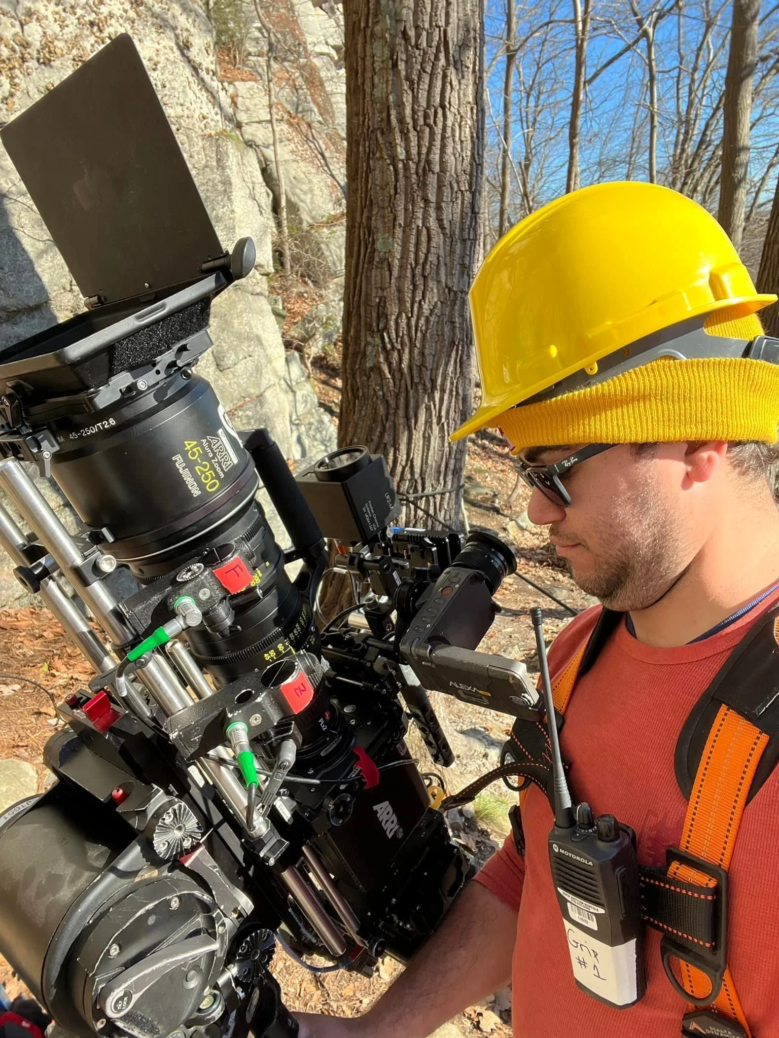A person in an orange shirt wearing a yellow hard hat is operating an ARRI film camera outdoors. They have a walkie-talkie attached to their harness, standing near trees and rocks.