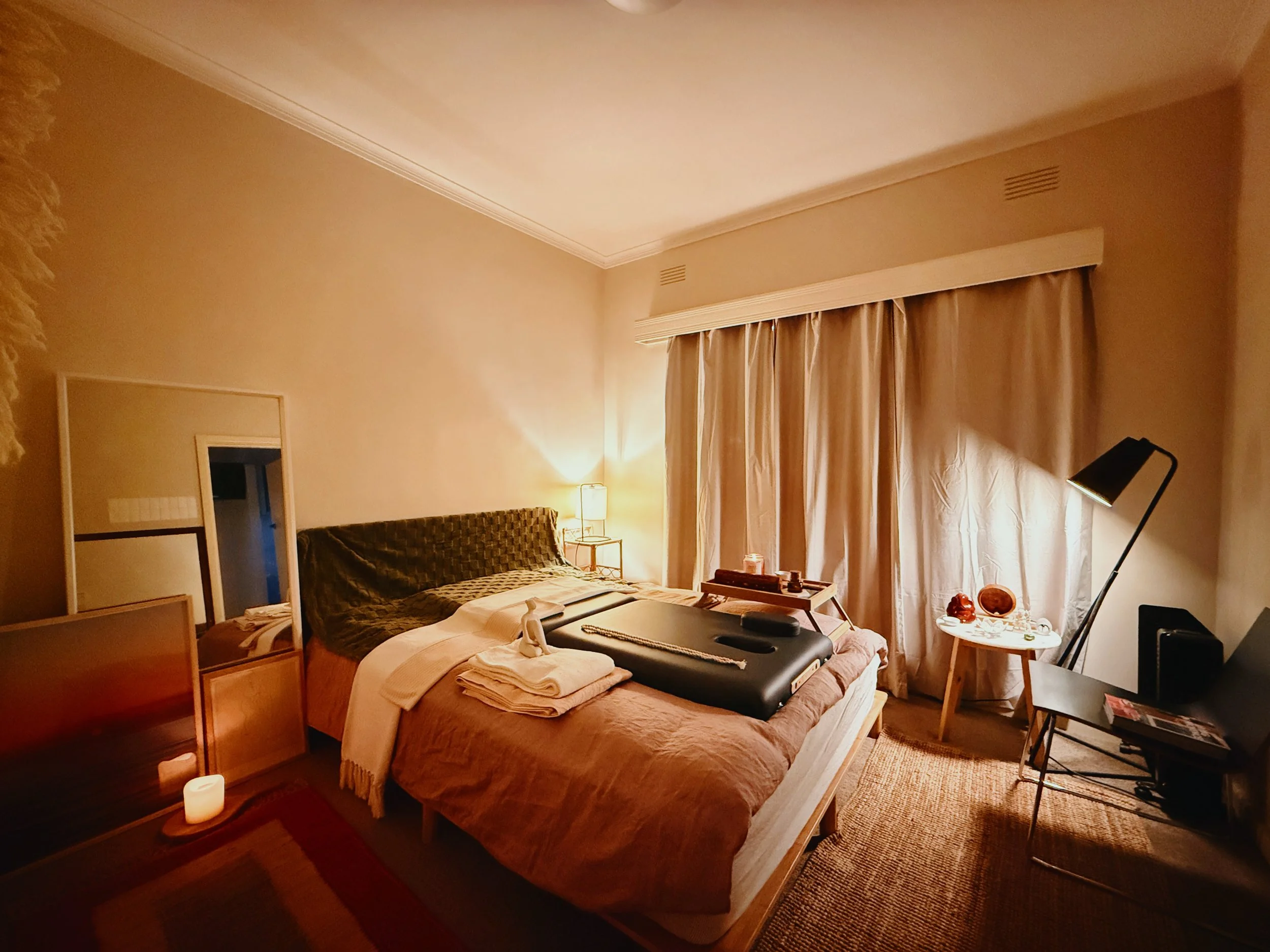 Cozy bedroom with warm lighting, a bed with pillows and blankets, a mirror on the floor, and a bedside table with a lamp and candles. Curtains cover the window, and the room has a relaxed, inviting atmosphere.