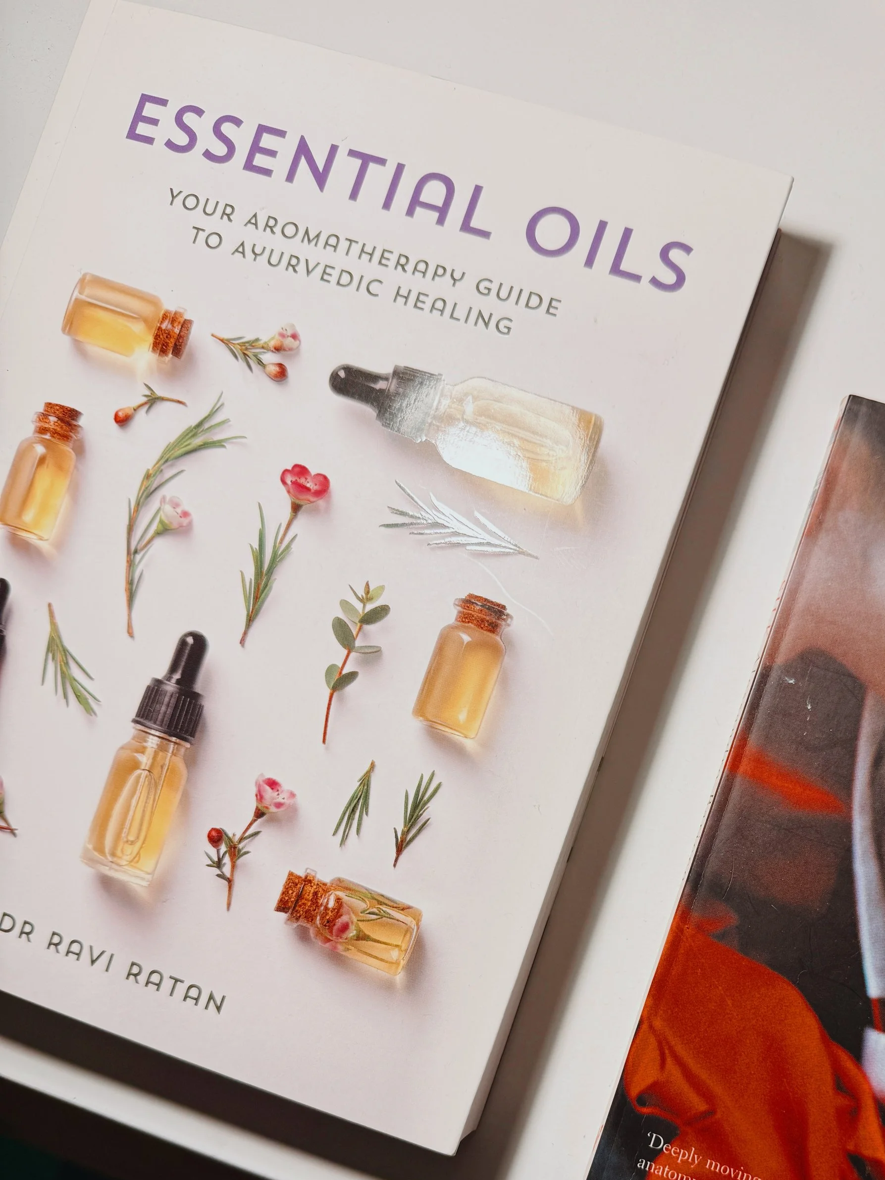Books on a white surface: 'Essential Oils' by Dr. Ravi Ratan with images of oils and herbs on the cover, and 'Silk' by Alessandro Baricco with a partial image of a person in red and white clothing.