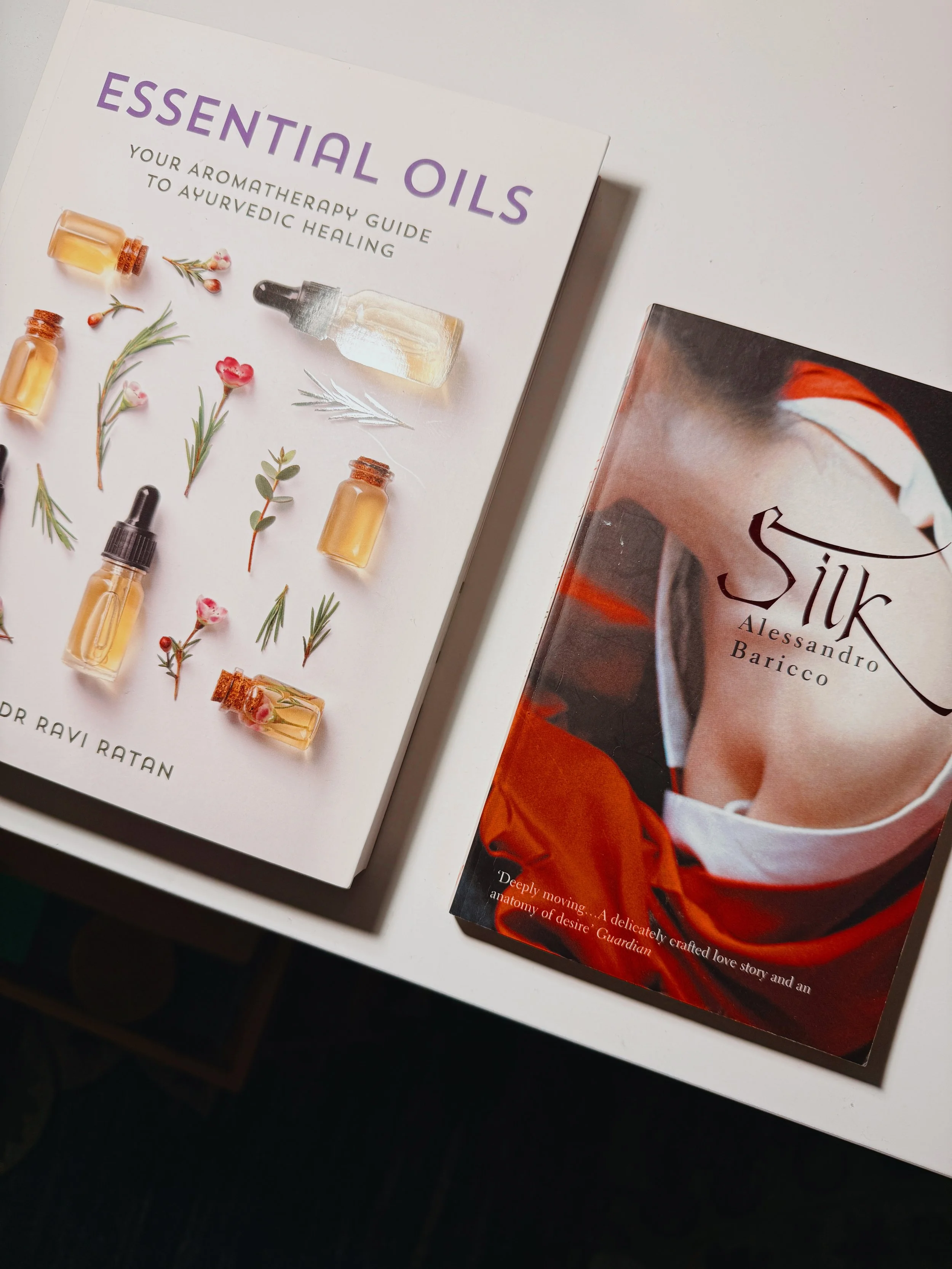 Books on a white surface: 'Essential Oils' by Dr. Ravi Ratan with images of oils and herbs on the cover, and 'Silk' by Alessandro Baricco with a partial image of a person in red and white clothing.