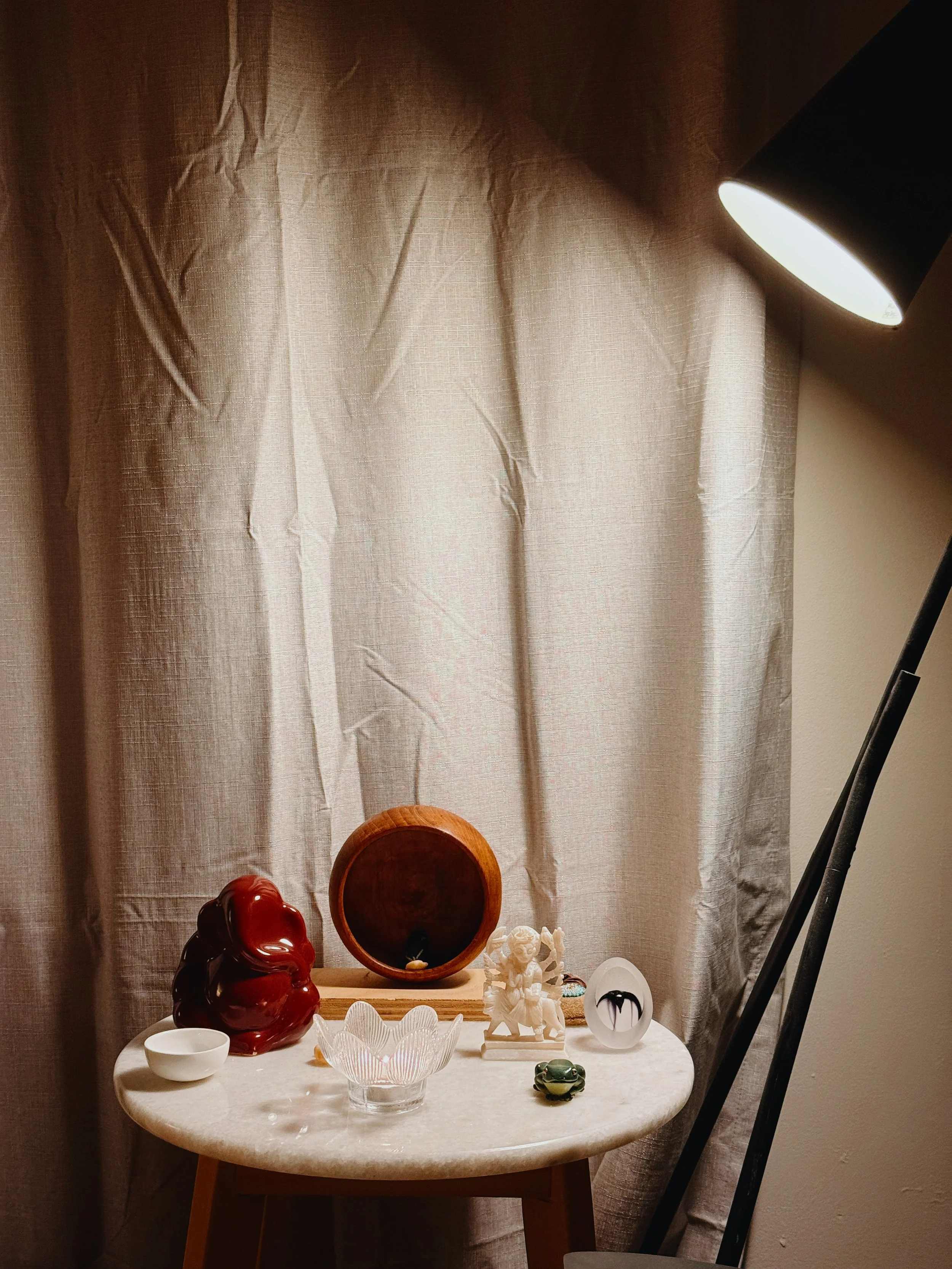A small round table with various decorative items including a red sculpture, a white figurine, a glass bowl, and other small objects, in front of a beige curtain under a floor lamp.