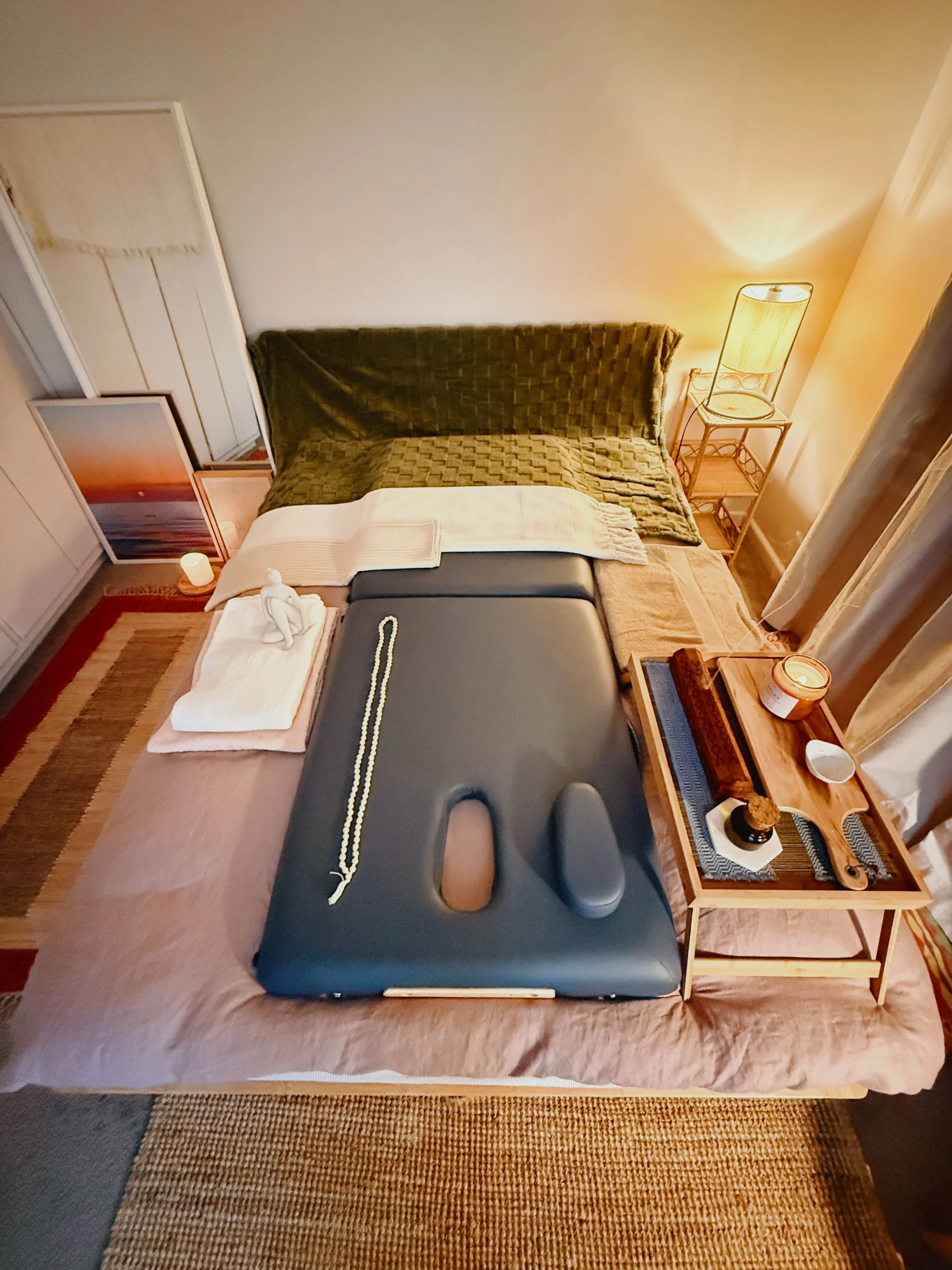 Cozy spa room with massage table, green blanket, framed artwork, lamp, and candle.