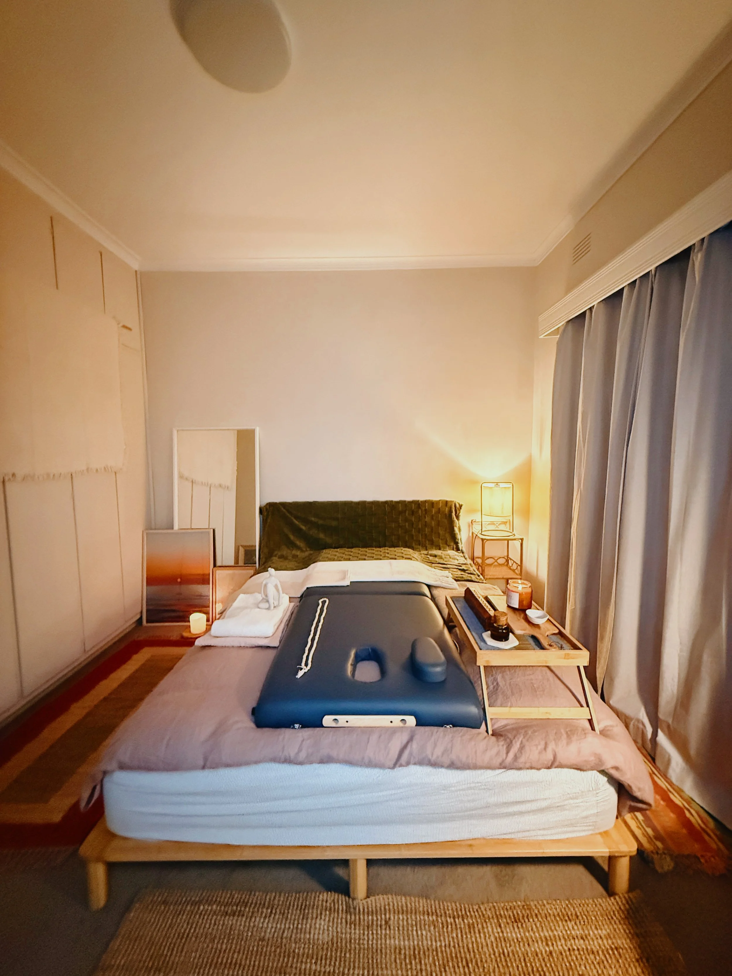 A cozy bedroom with a neatly made bed featuring a padded headboard against the wall. The bed has a small wooden tray with a candle and a cup on it. A cushiony blue mat is placed on the bed for relaxation. Curtains frame a large window, and a warm lam