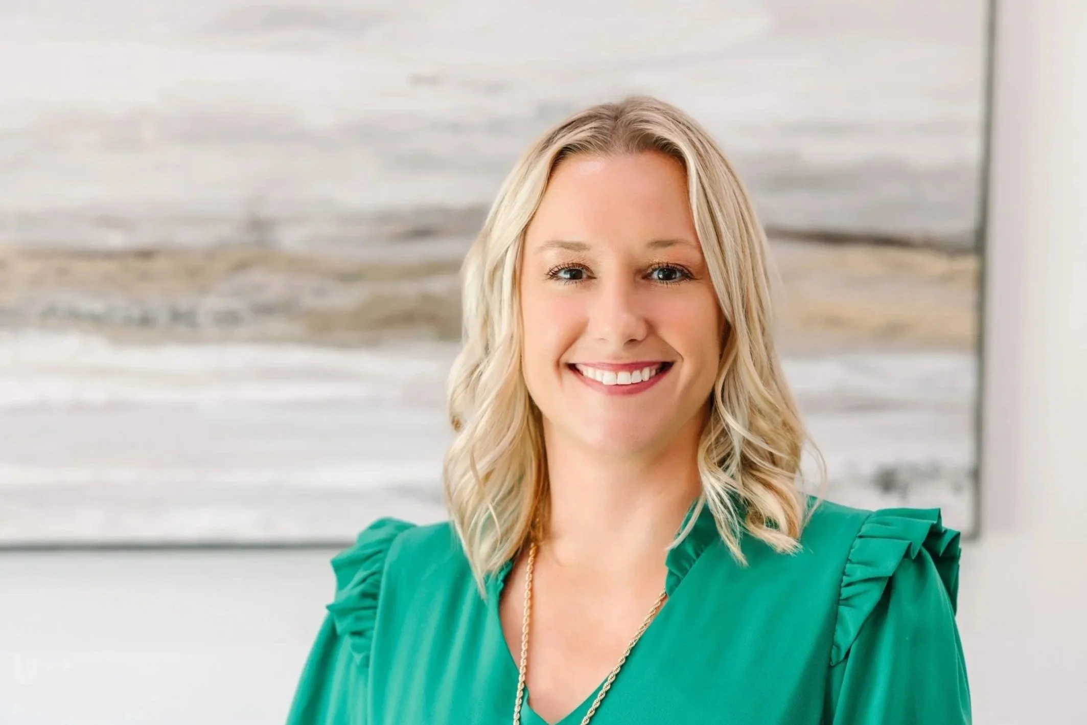 A woman with blonde, wavy hair smiling at the camera, wearing a green blouse with ruffled shoulders, in front of a coastal background.