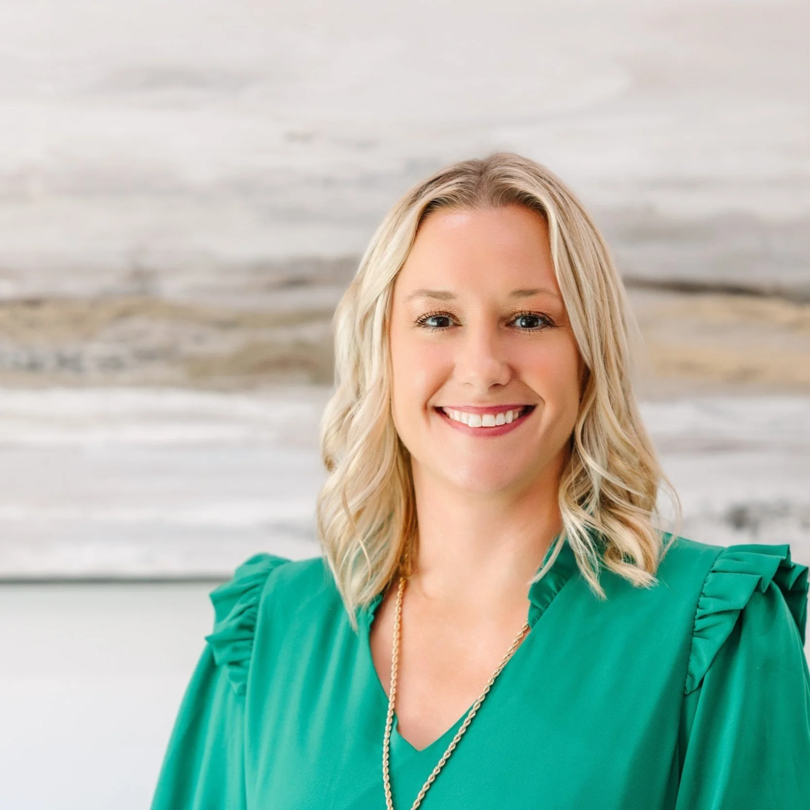 A woman with blonde hair, smiling, wearing a green blouse with ruffled sleeves, standing in front of a large abstract painting with neutral tones.
