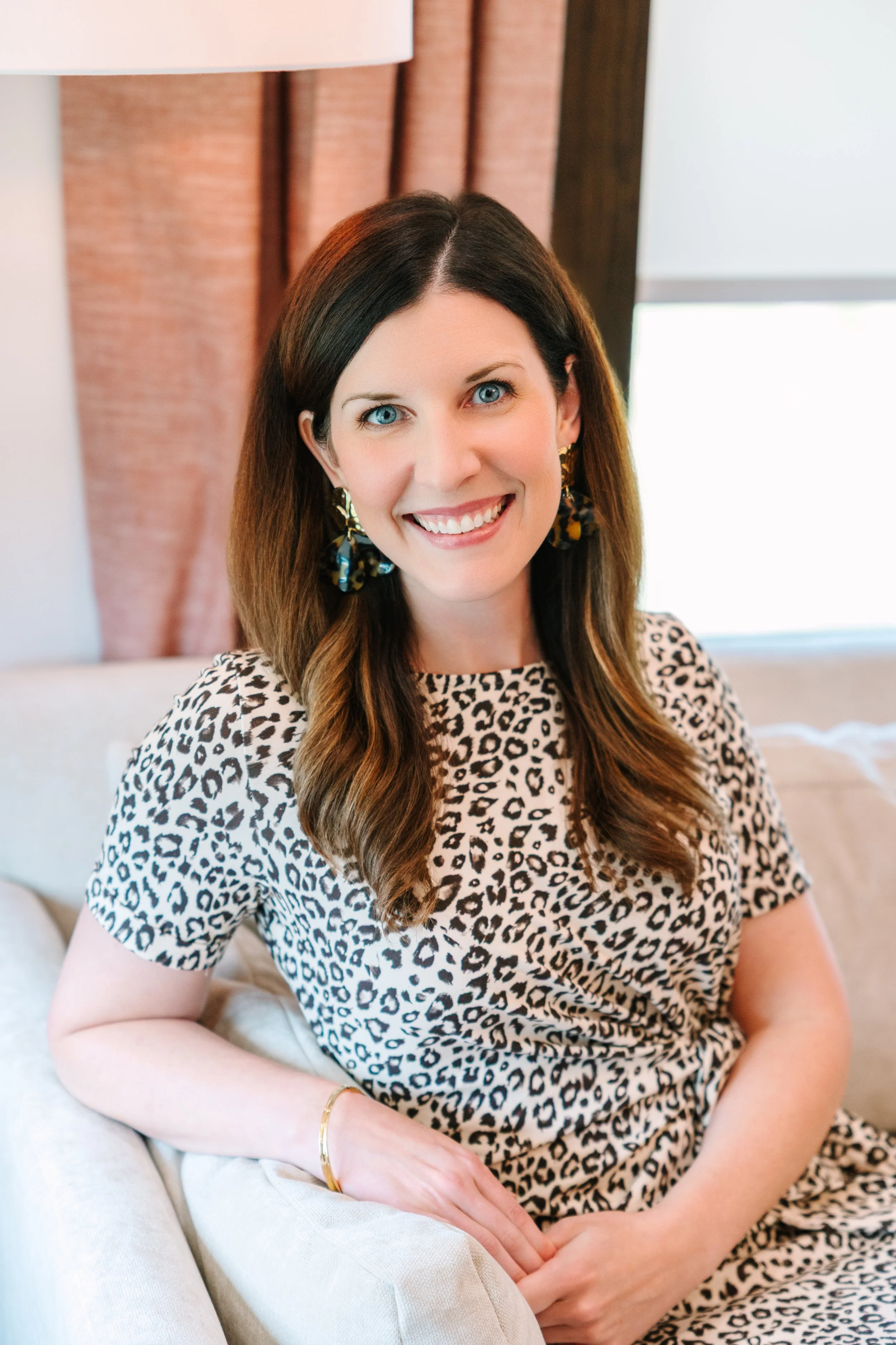 A woman with long brown hair, wearing a leopard print dress and large earrings, smiling while sitting on a beige sofa in a room with curtains and a window.