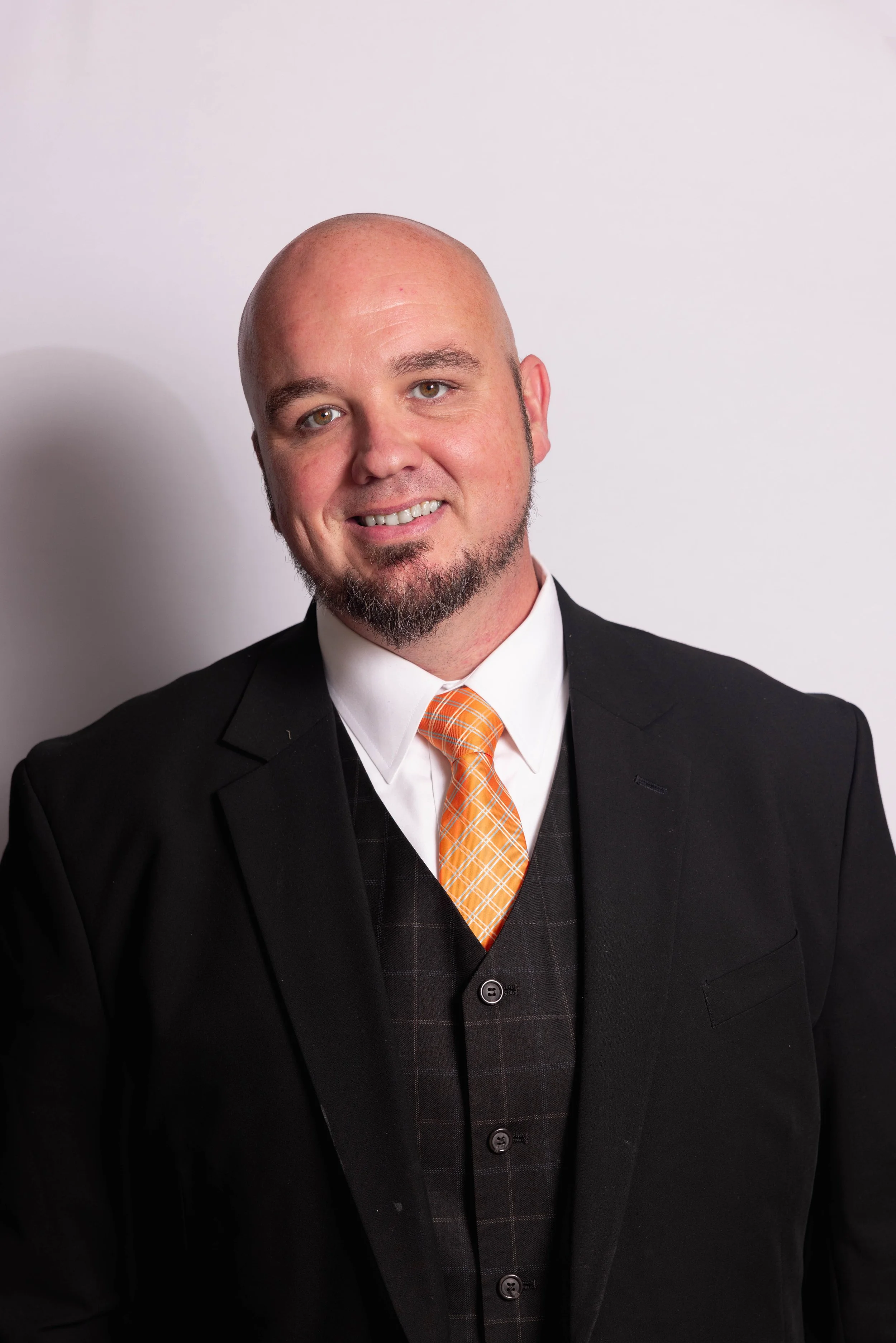 A man with a bald head, goatee, and mustache wearing a black suit jacket, white shirt, and orange checkered tie, smiling against a plain white background.
