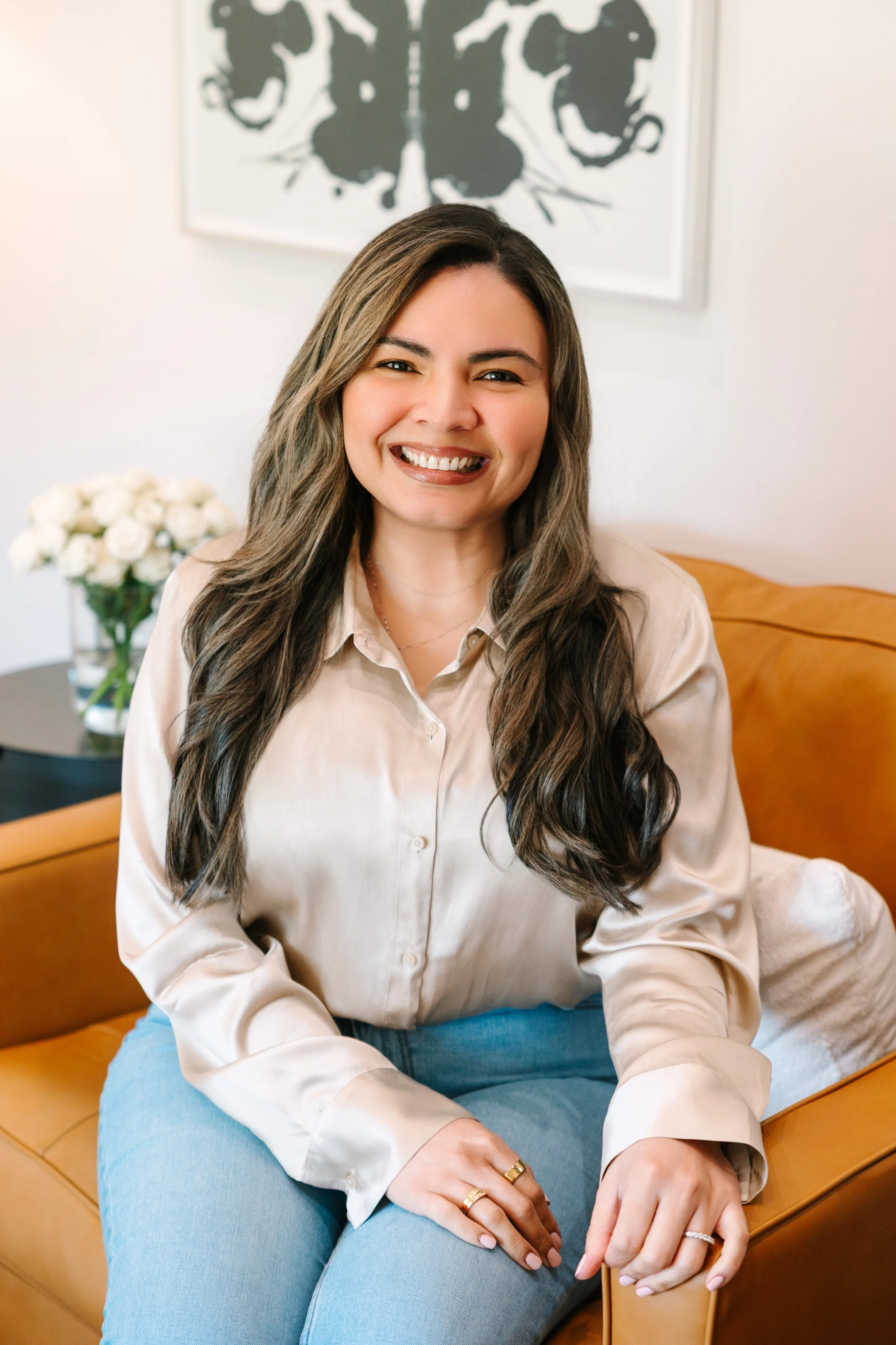 A woman with long wavy brown hair smiling while sitting on a tan leather couch in a well-lit room. She is wearing a beige button-up shirt and light blue jeans, with multiple rings on her fingers. In the background, there is a white wall with a black and white butterfly artwork and a side table with a vase of white roses.