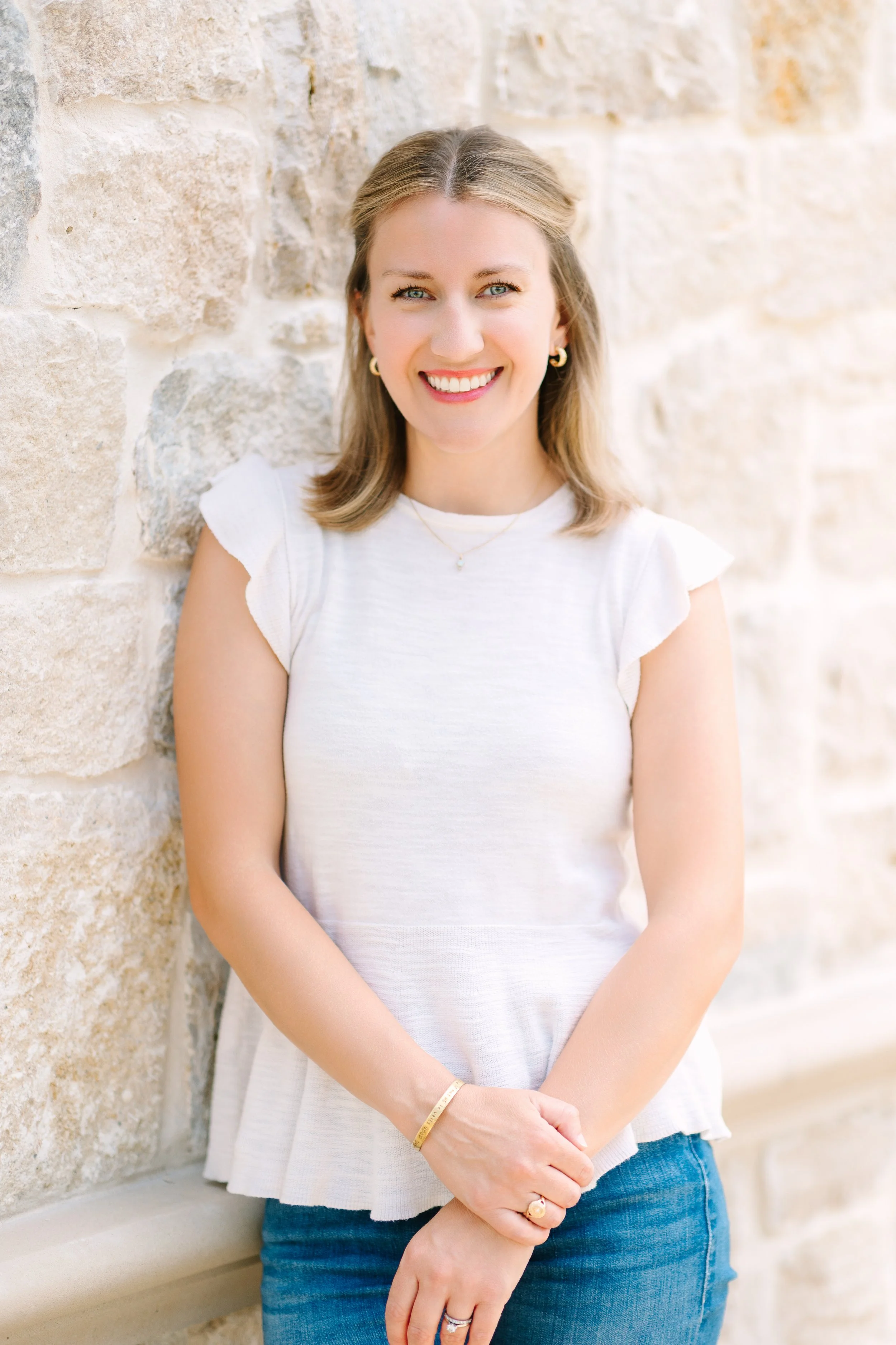 A young woman with blonde hair, blue eyes, and a bright smile, standing against a stone wall, wearing a white short-sleeved top and blue jeans.