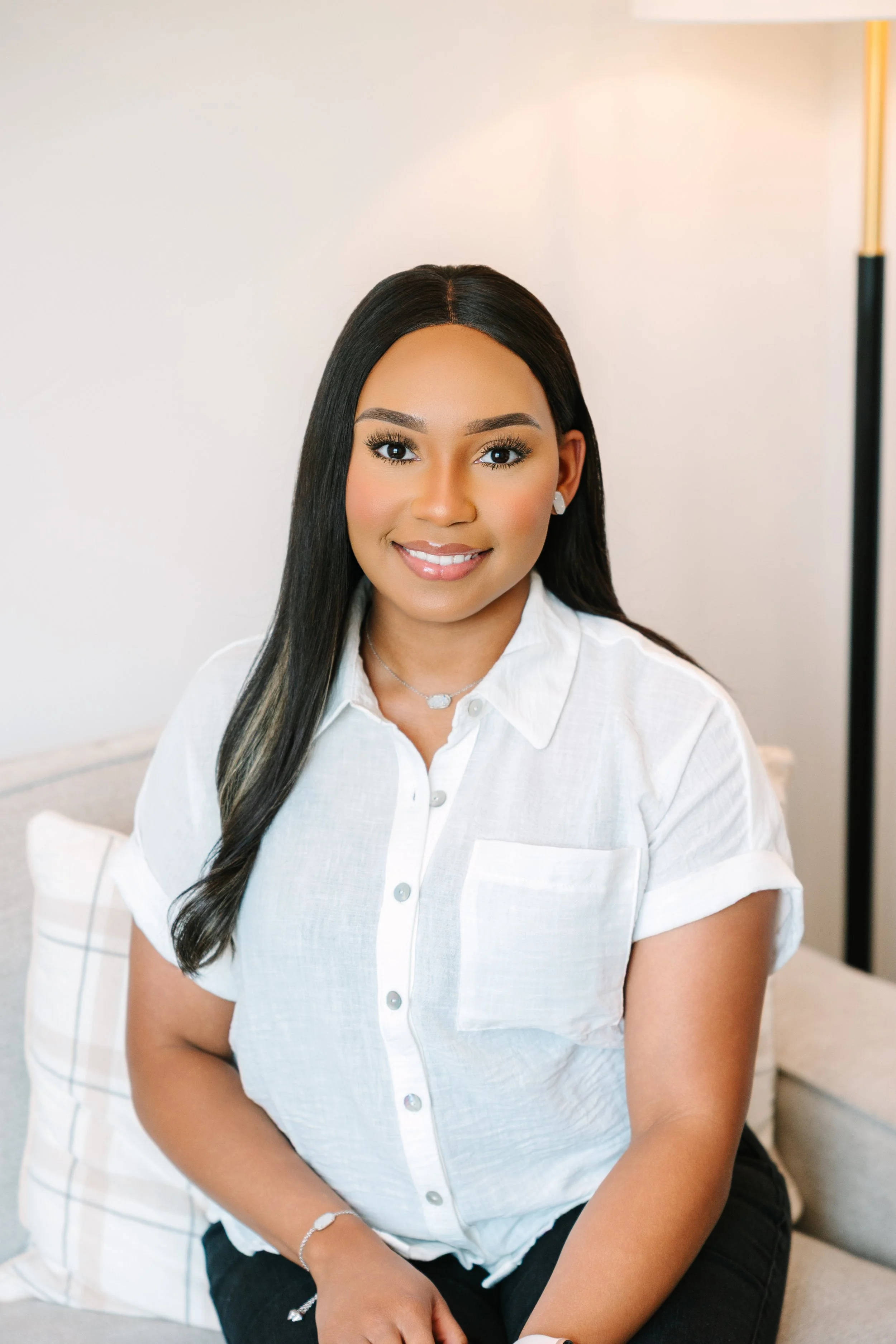 A young woman with long black hair and a warm smile, wearing a white button-up shirt and sitting on a beige couch with pillows, in a bright, well-lit room.