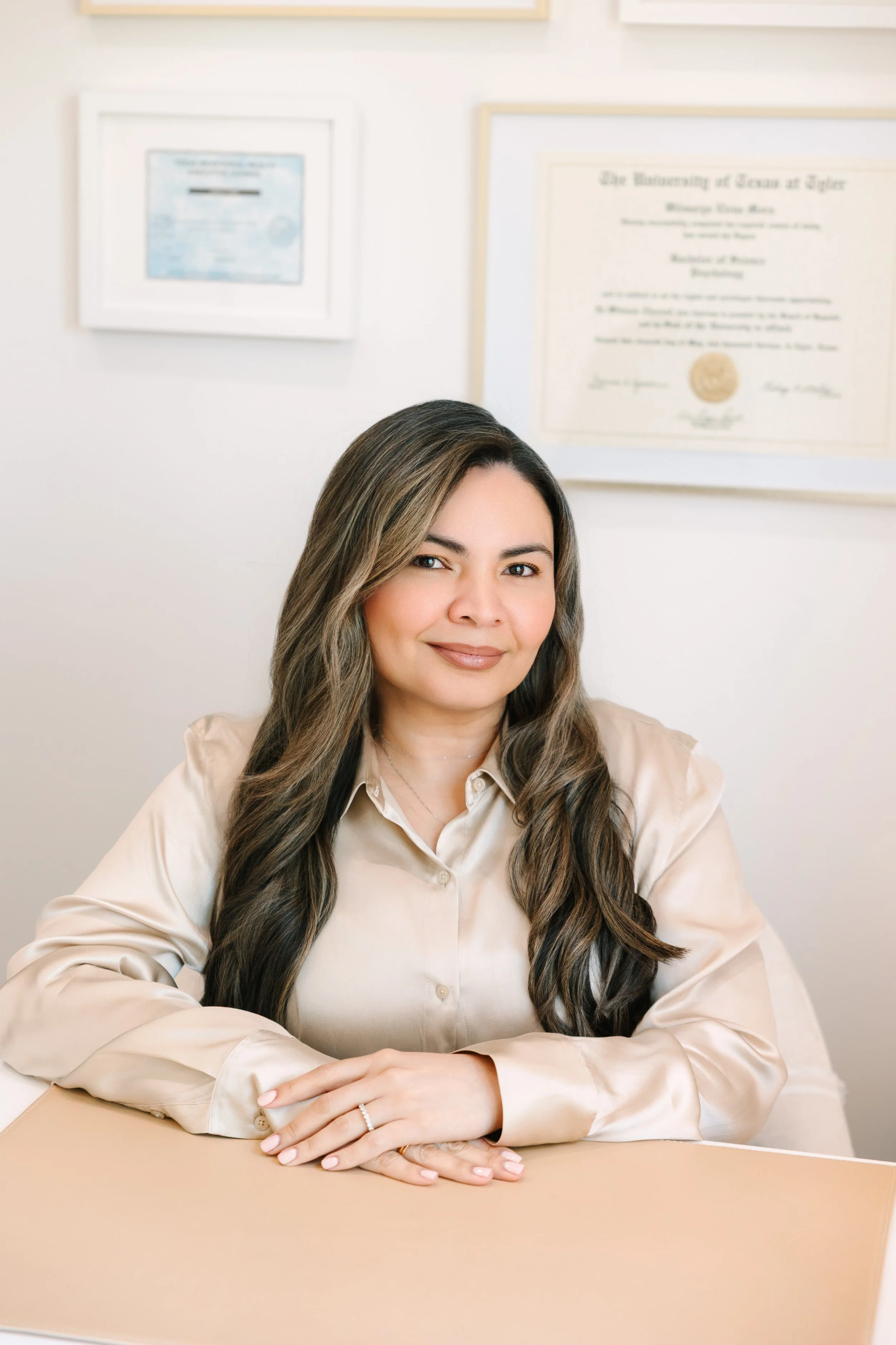 A woman with long wavy brown hair smiling while sitting on a tan leather couch in a well-lit room. She is wearing a beige button-up shirt and light blue jeans, with multiple rings on her fingers. In the background, there is a white wall with a black and white butterfly artwork and a side table with a vase of white roses.
