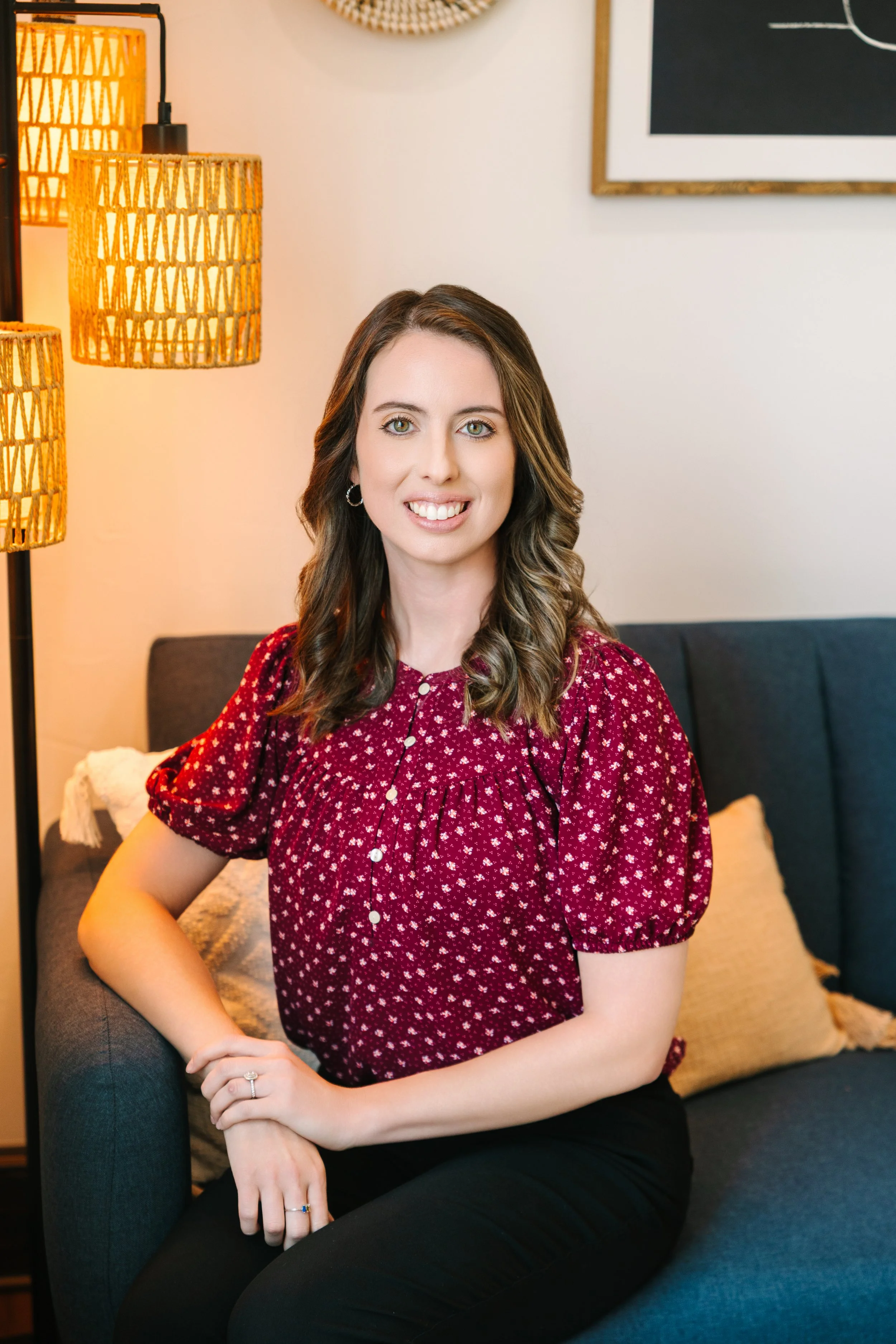 A young woman with shoulder-length wavy brown hair, wearing a red patterned blouse, sitting on a dark blue sofa with a beige pillow in a cozy, well-lit room with warm lighting, a black framed chalkboard, and wicker lamps in the background.