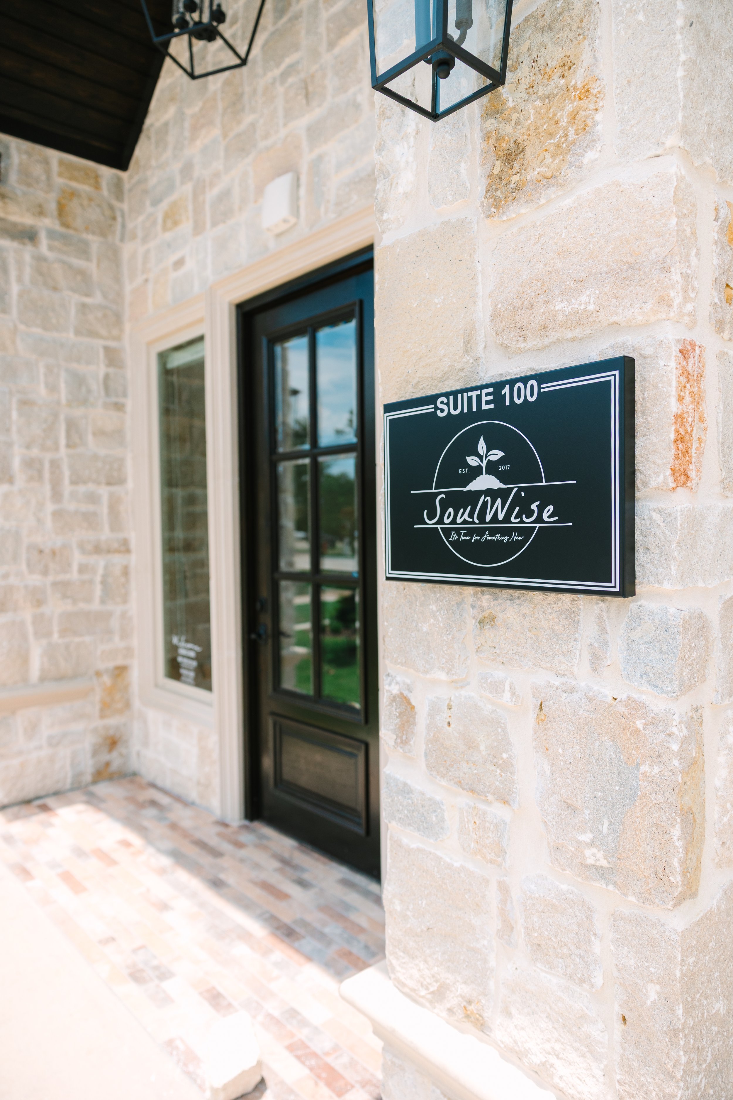 Exterior of a building with a black door and stone wall, showing a black sign reading "Suite 100 SoulWise" with a slogan "Life Tips for Something New" and a logo of a sprouting plant.