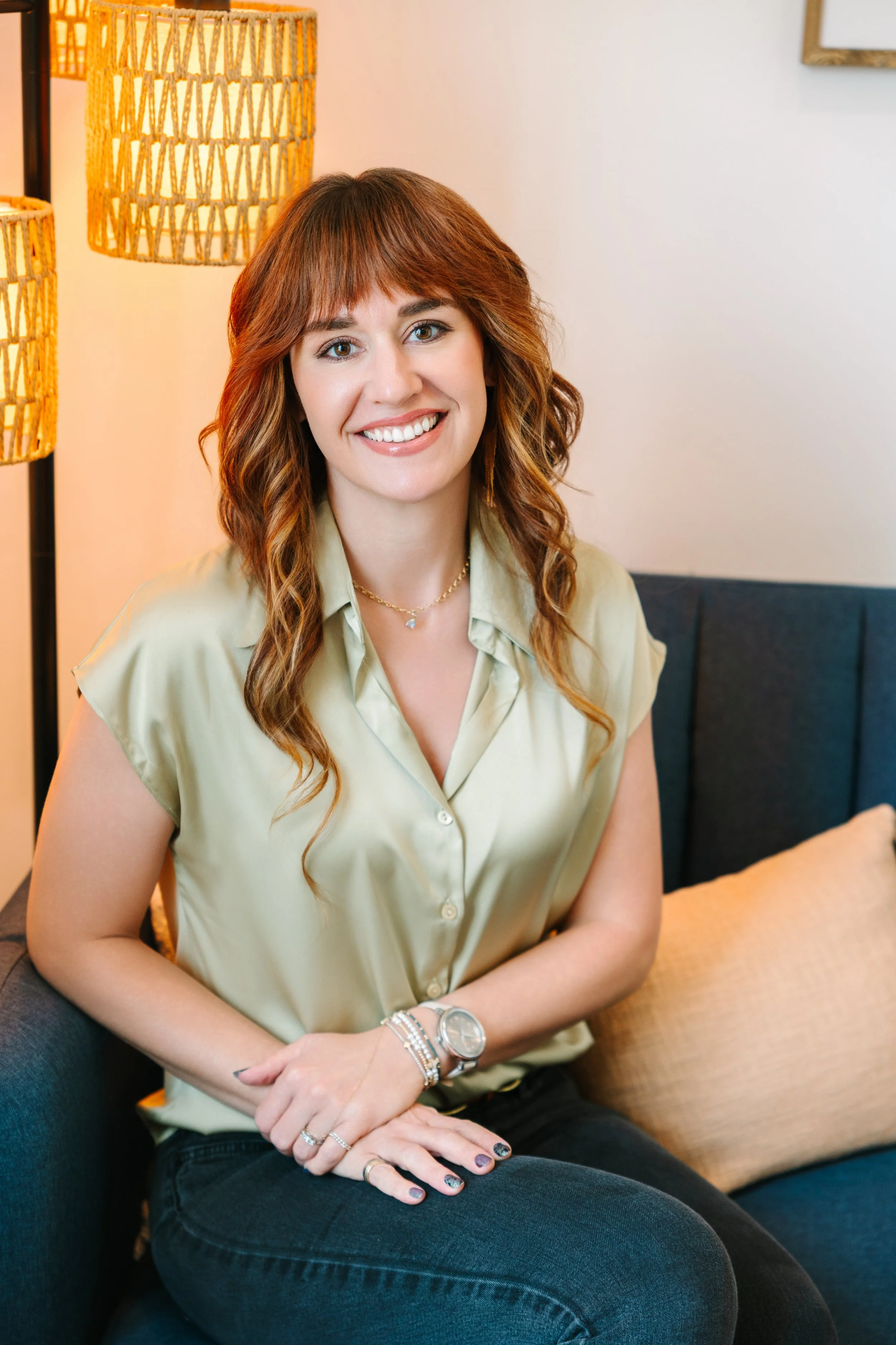 A woman with long, wavy auburn hair and light skin, sitting on a blue sofa in a cozy living room. She is smiling, wearing a light green satin blouse, several bracelets, a watch, and rings. Behind her are warm-colored lamps and a pillow.