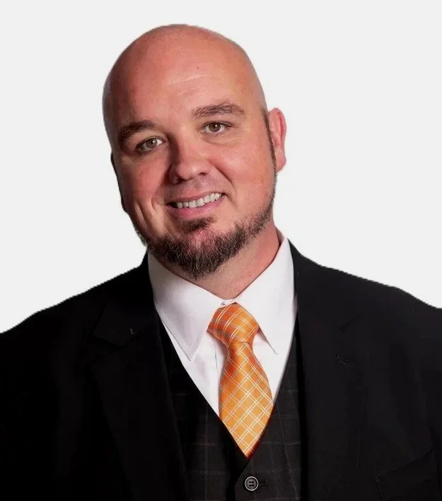 A man with a bald head, goatee, and mustache wearing a black suit jacket, white shirt, and orange checkered tie, smiling against a plain white background.