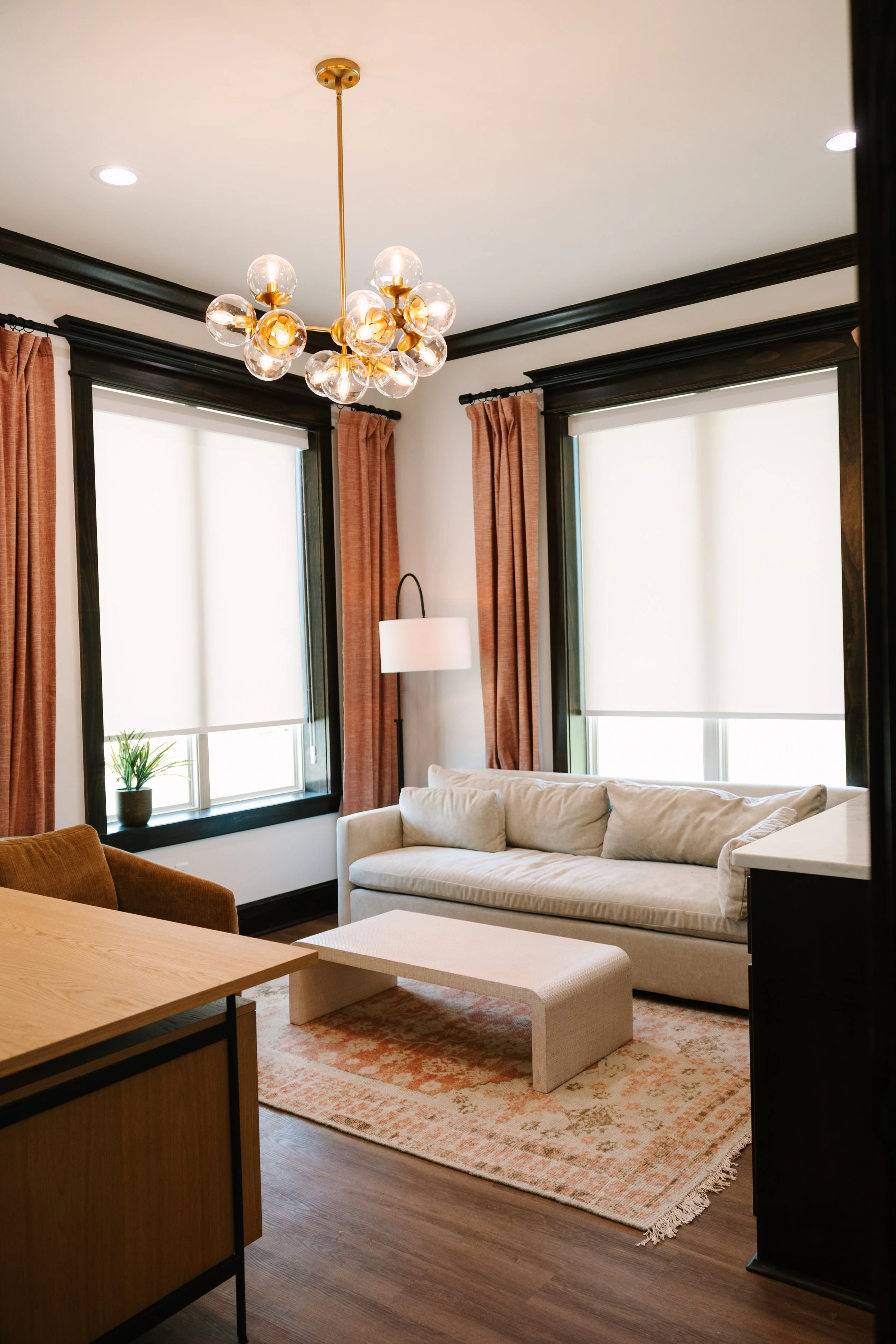 Living room with white couch, beige coffee table, orange curtains, black window frames, and a modern ceiling light fixture.
