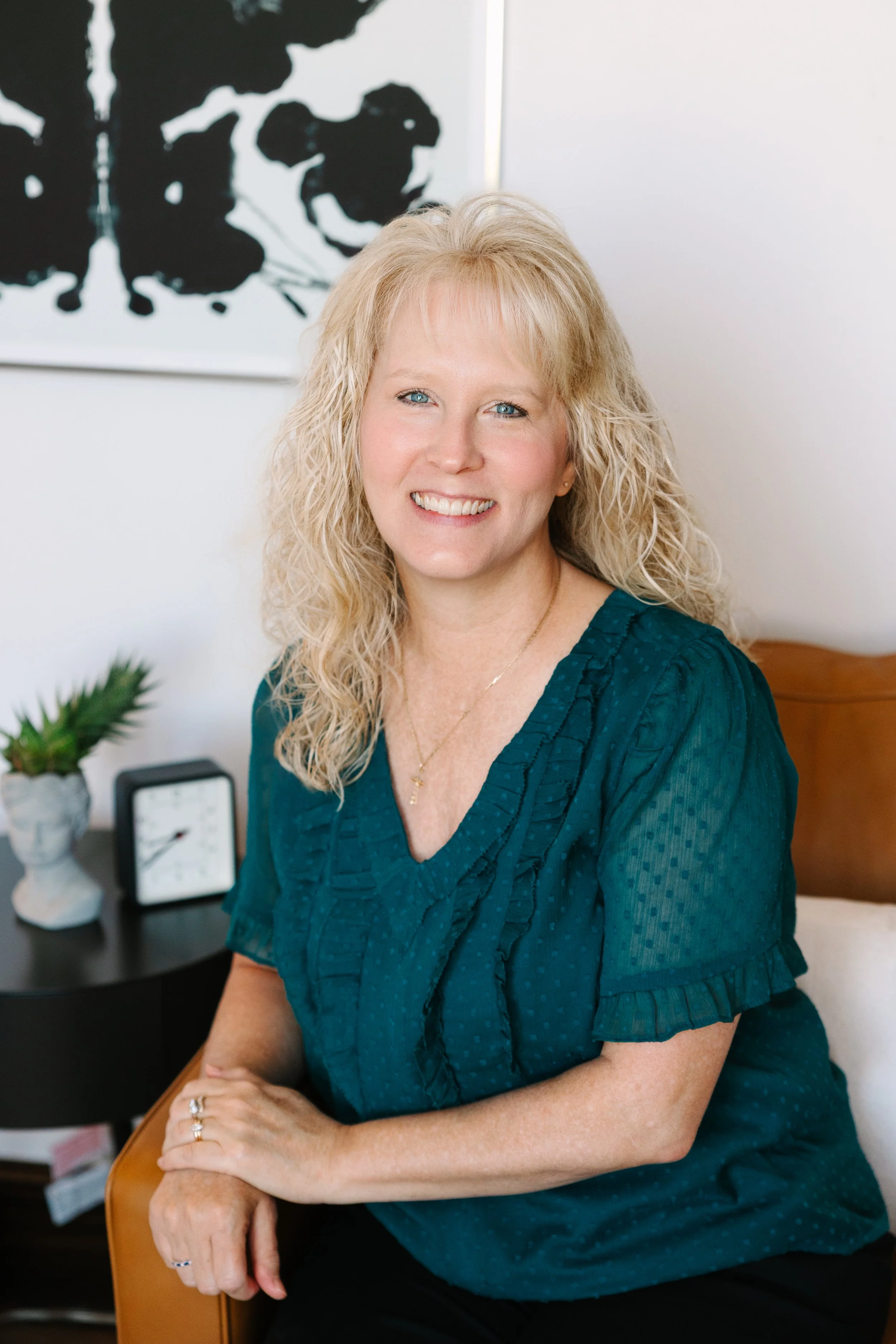 A woman with blonde curly hair, smiling, sitting on a chair in a room with white walls, wearing a teal blouse. There is a small black clock and a potted plant on a dark bedside table behind her, with a black and white abstract painting on the wall.